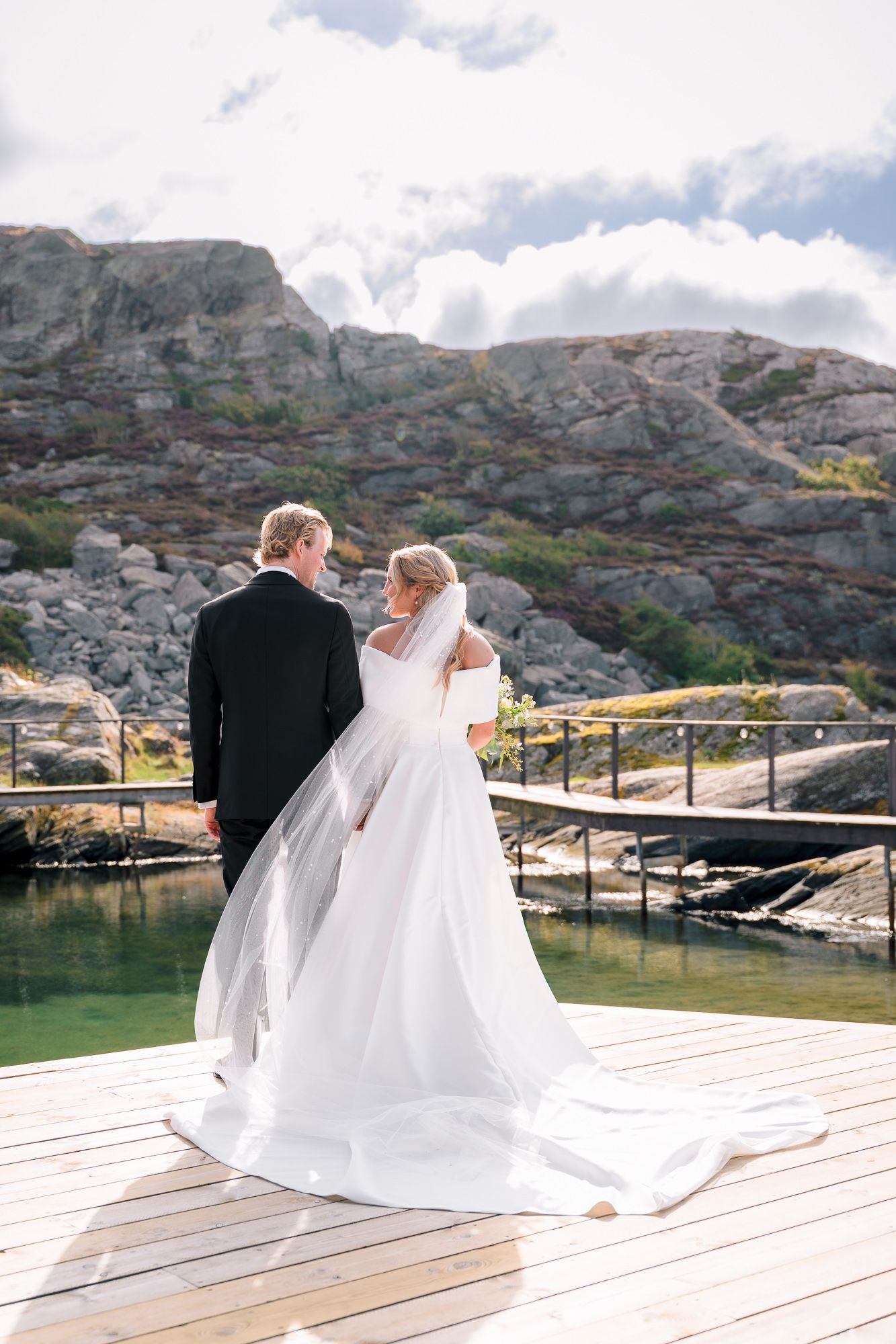 A couple in wedding attire stands on a wooden dock overlooking a scenic mountain lake on their special day.