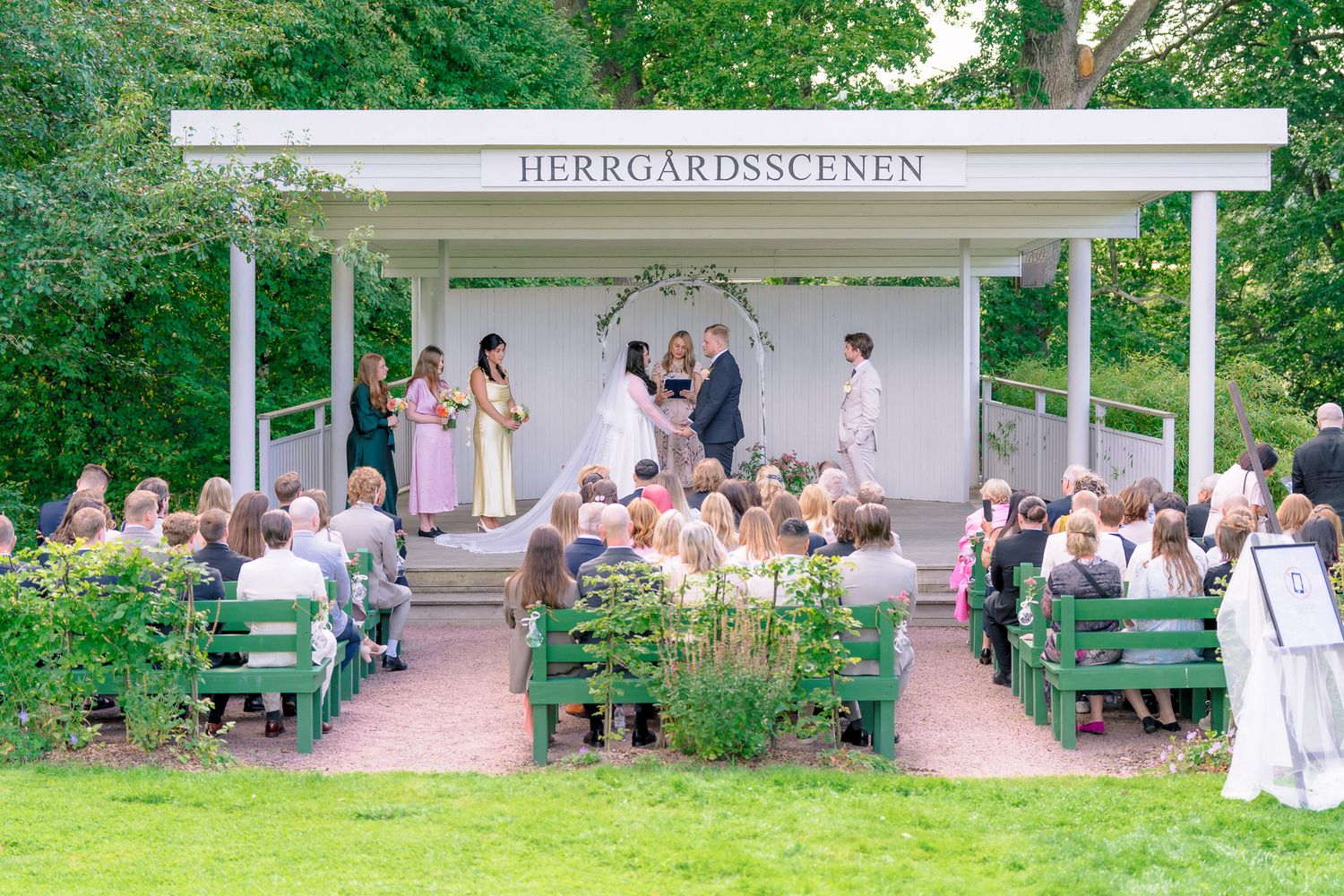 An outdoor wedding ceremony takes place at a white pergola venue surrounded by lush gardens and seated guests.