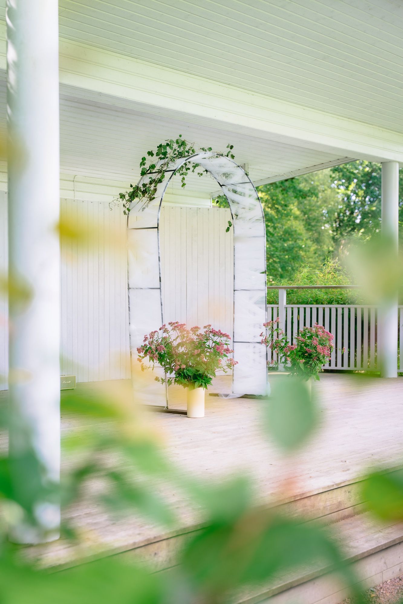 White farmhouse porch with classic columns and railing is framed by soft green leaves in a dreamy summer scene.
