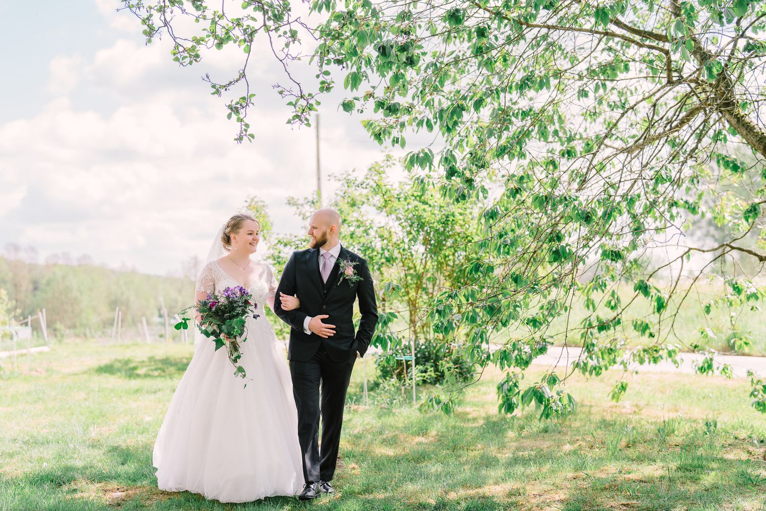 A wedding couple walks together on a grassy hill beneath blooming spring trees on a bright cloudy day.