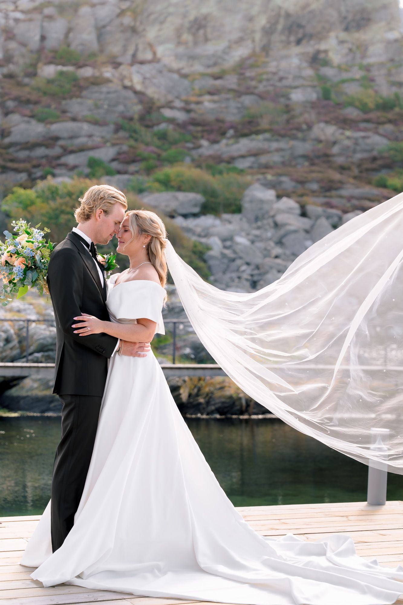 A romantic wedding couple embraces on a dock by rocky cliffs as the bride's long veil flows dramatically in the wind.