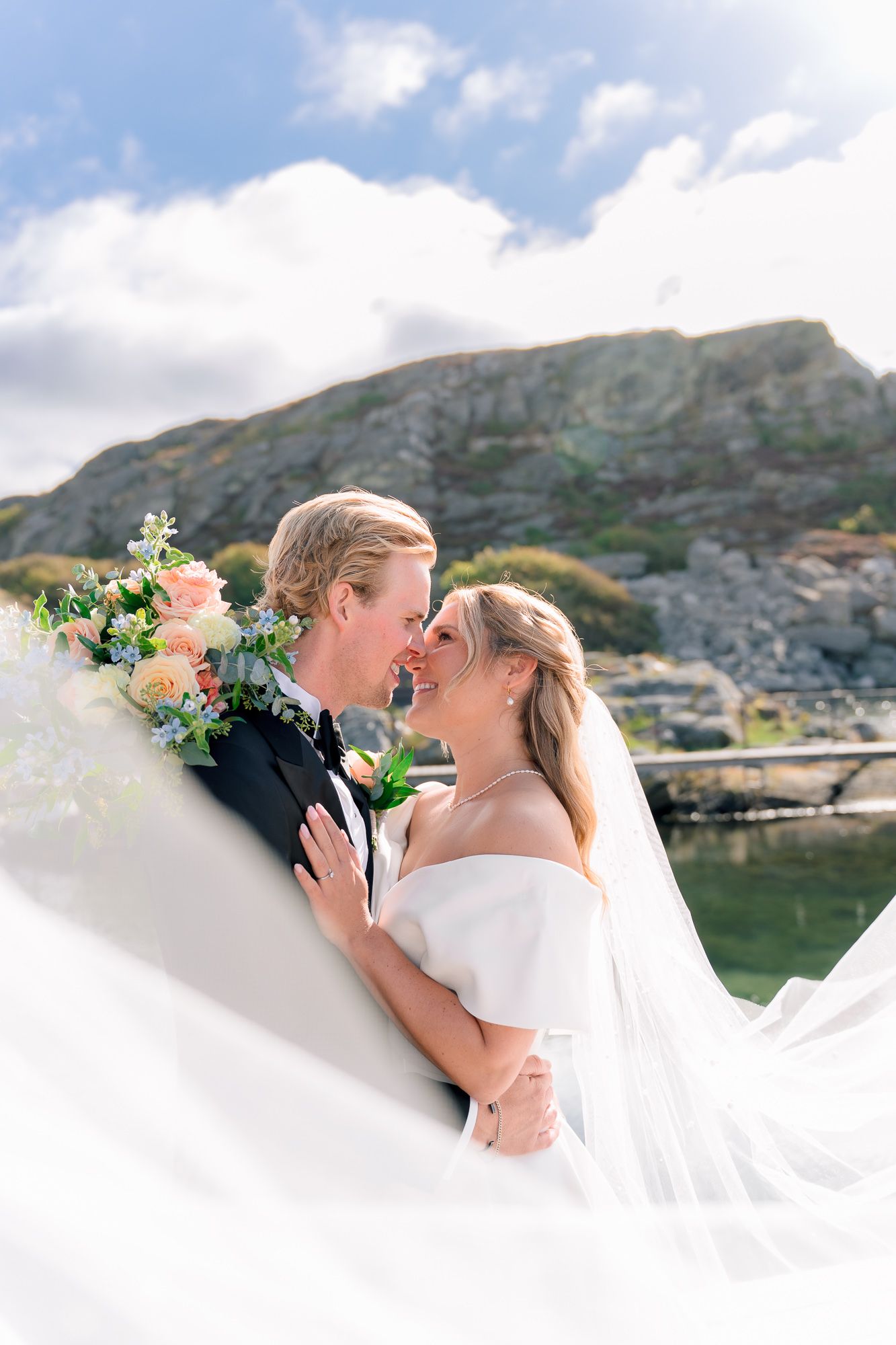 A romantic wedding couple shares an intimate moment against a scenic mountain backdrop with flowing white veil and floral bouquet.