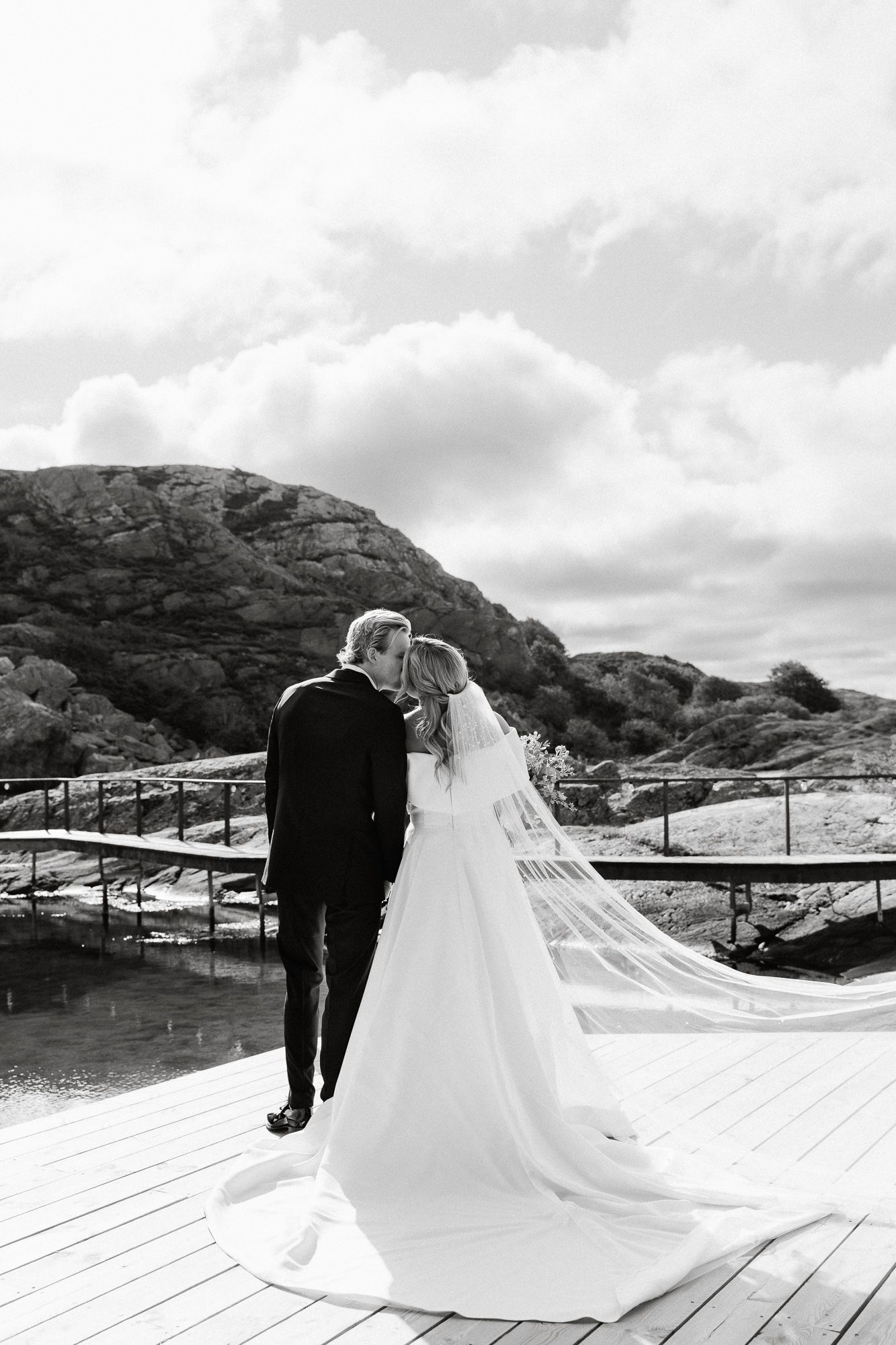 Panoramic wedding scene with couple in formal attire standing on wooden deck against mountainous backdrop.