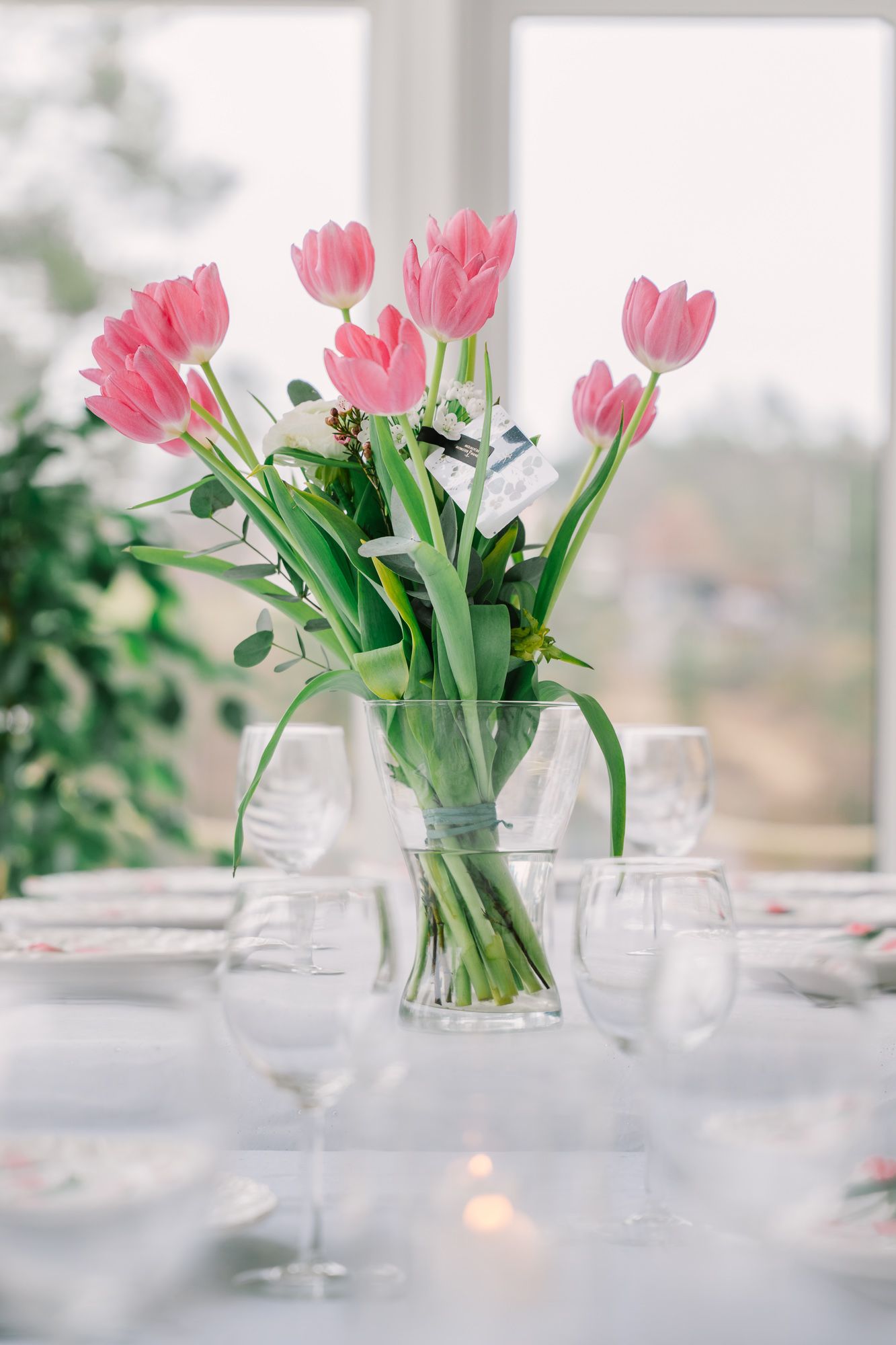 Pink tulips in a clear glass vase centerpiece surrounded by wine glasses on white tablecloth.