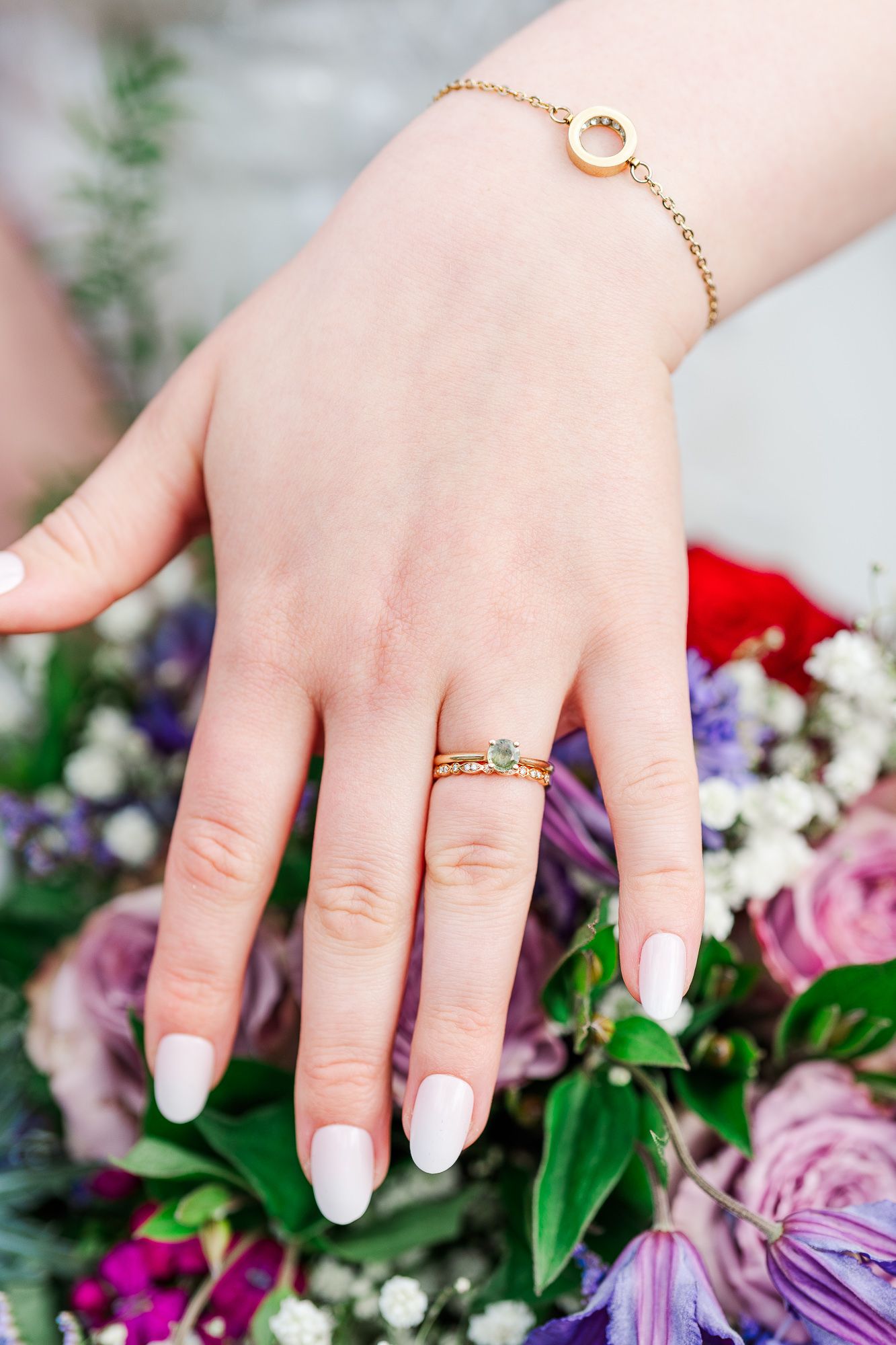 Delicate hand wearing white nail polish, gold bracelet and rings rests on colorful floral bouquet with roses and purple flowers.