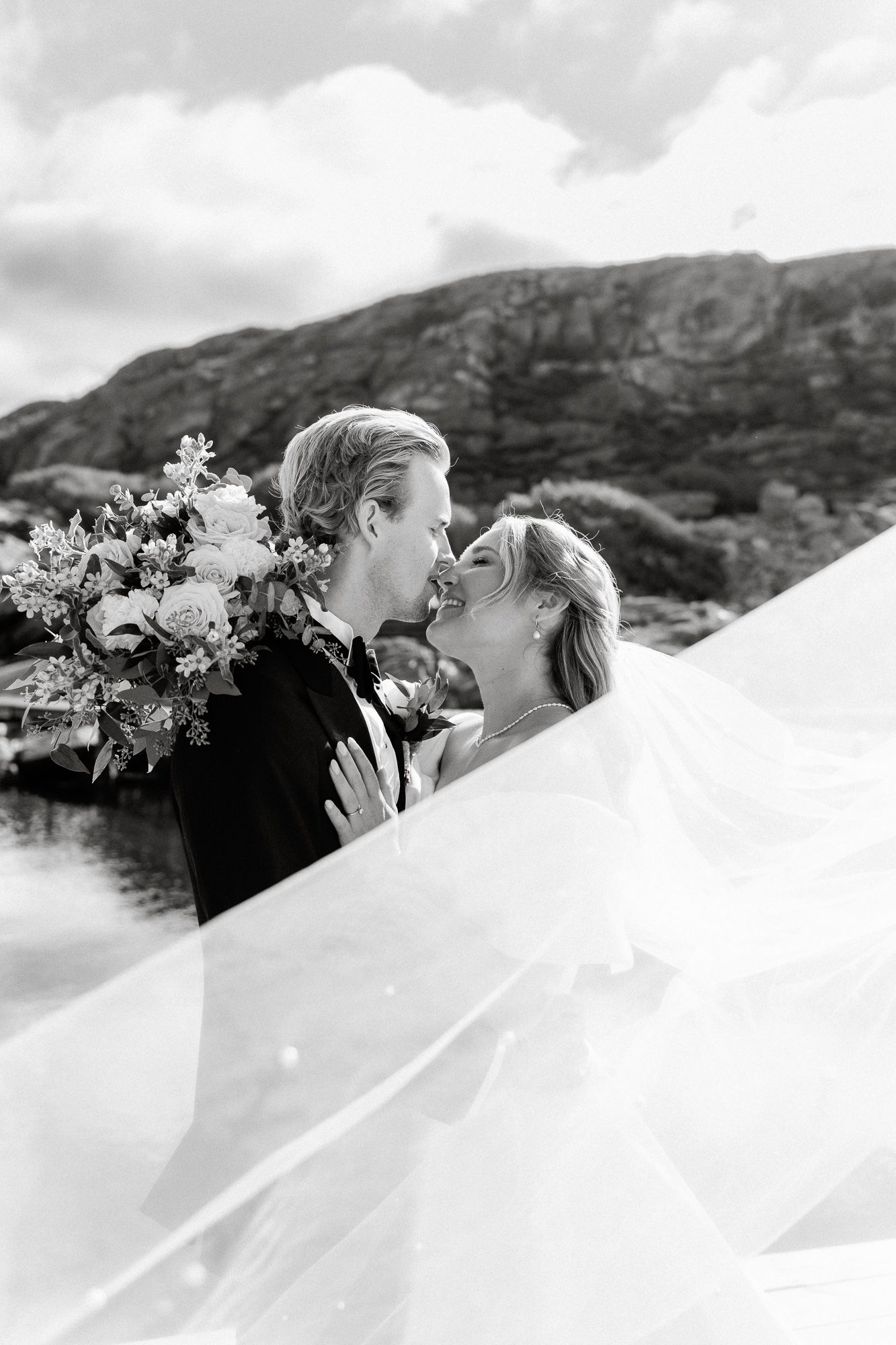 Black and white wedding photo captures a tender moment between newlyweds with mountains and dramatic veil in the background.