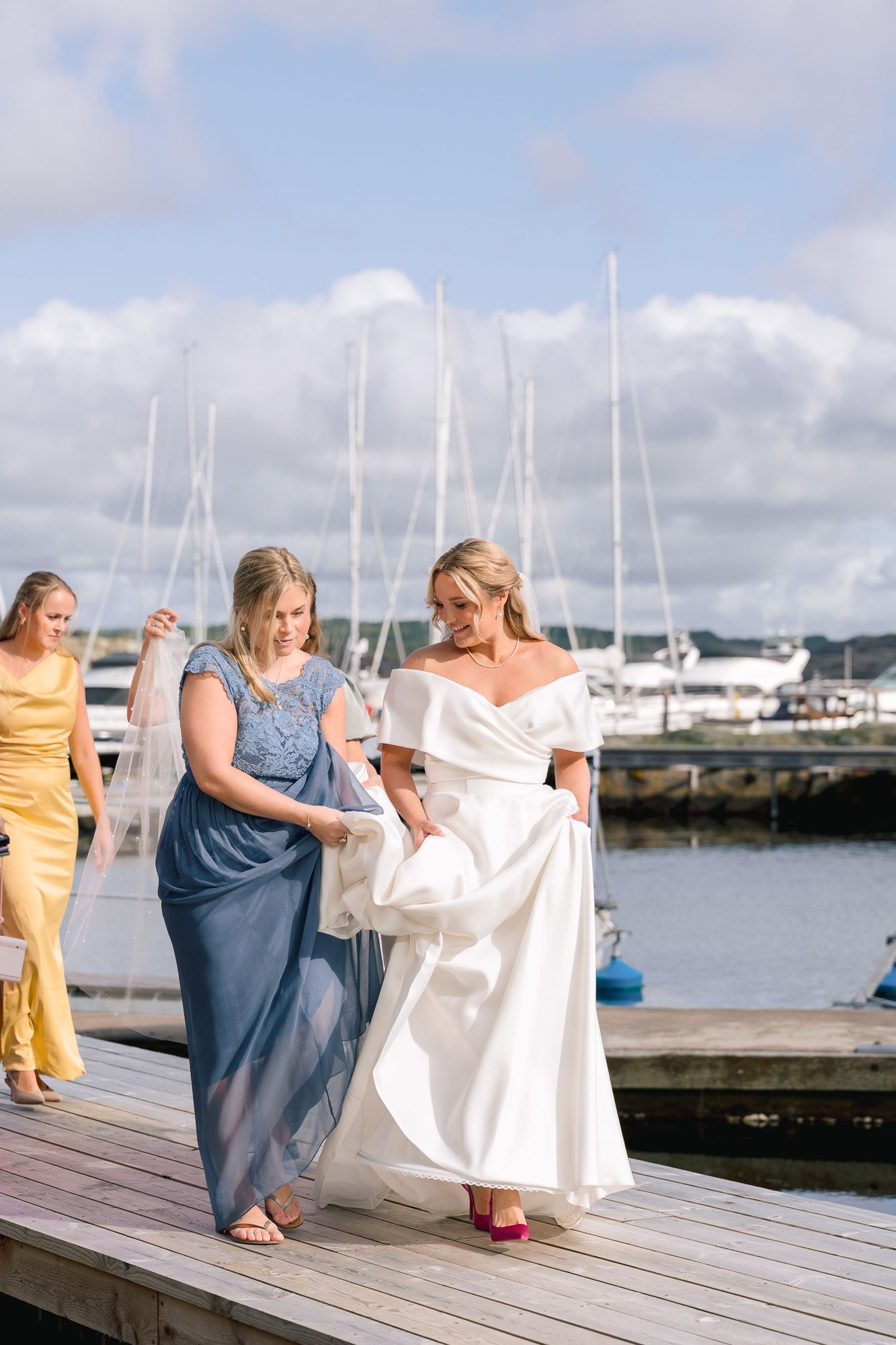 A group of women walking along a wooden dock at a marina with sailboats and cloudy skies in the background.