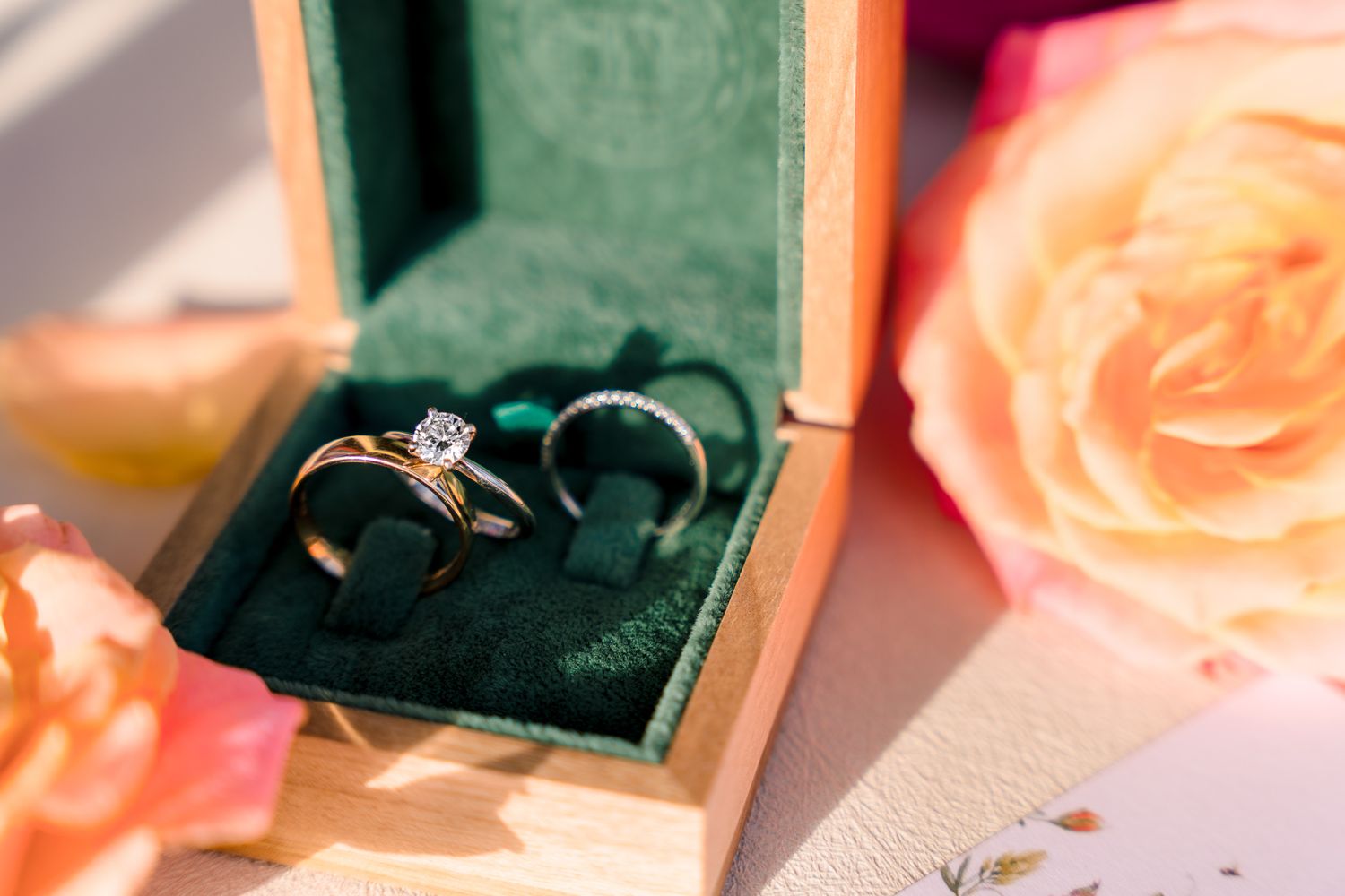 A sparkling diamond engagement ring sits nestled in an emerald green velvet ring box with peach roses in the background.