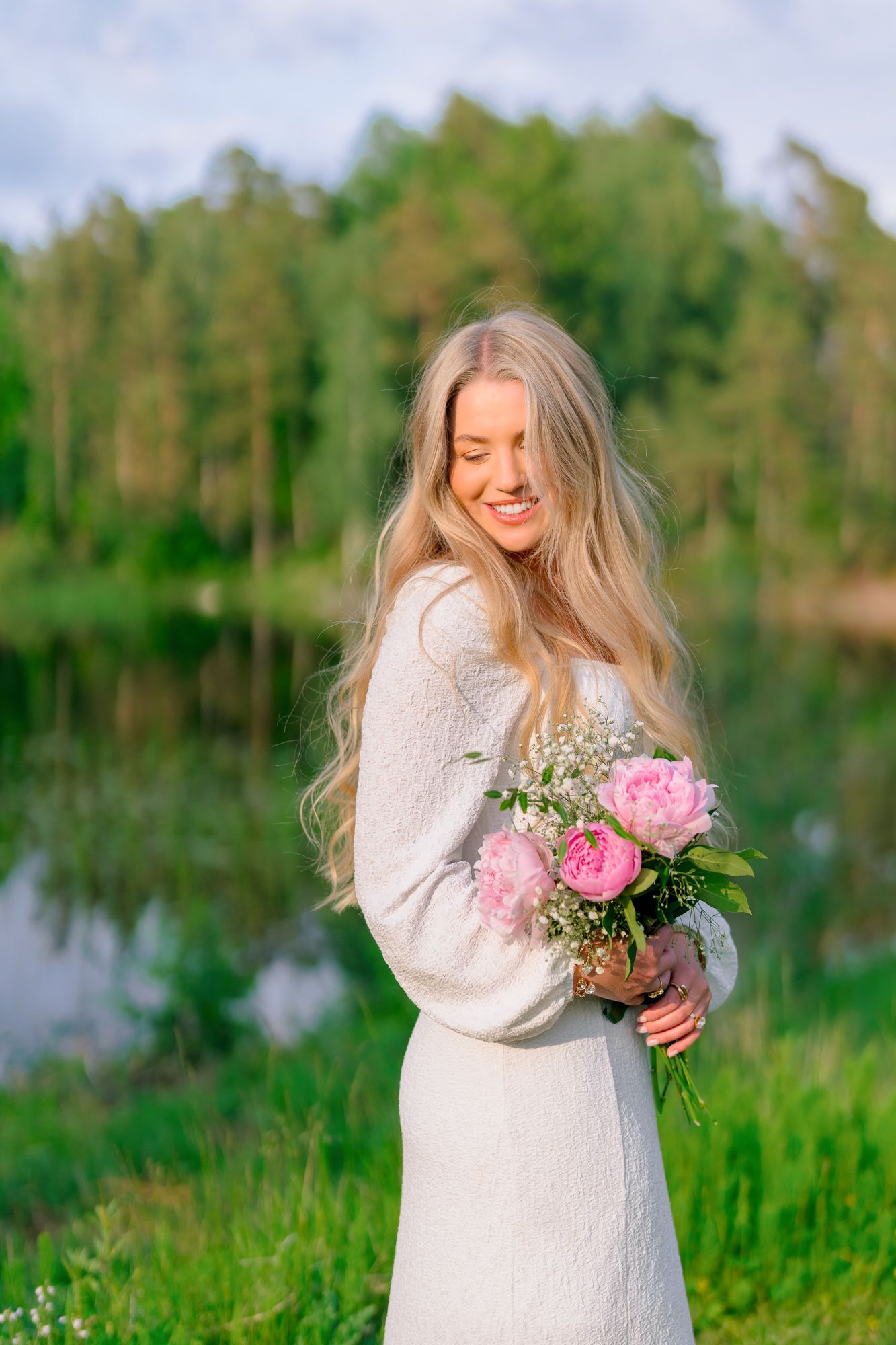 A person in a white sweater stands by a lake holding pink peonies against a green natural background.