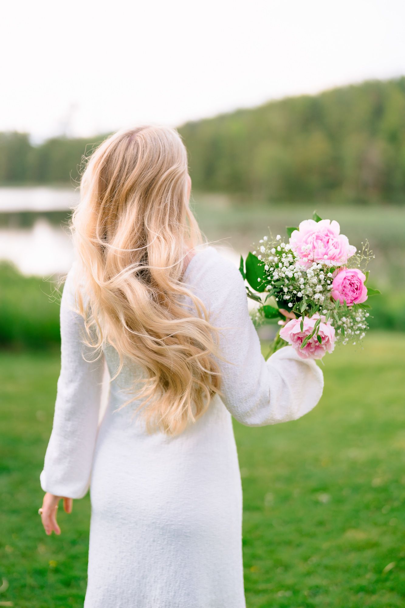 A person in a white sweater outdoors holding pink flowers with their blonde hair blowing in the wind, viewed from behind.