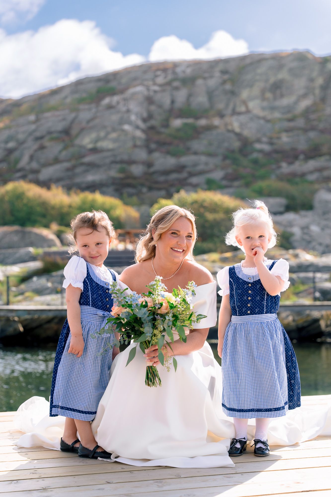 Young flower girls in blue dresses sit on wooden dock by mountain lake.