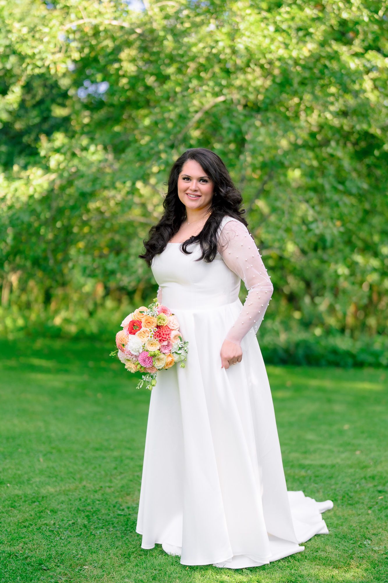 A bride in a white lace-sleeved wedding gown holds a pink and coral bouquet while standing on lush green grass.