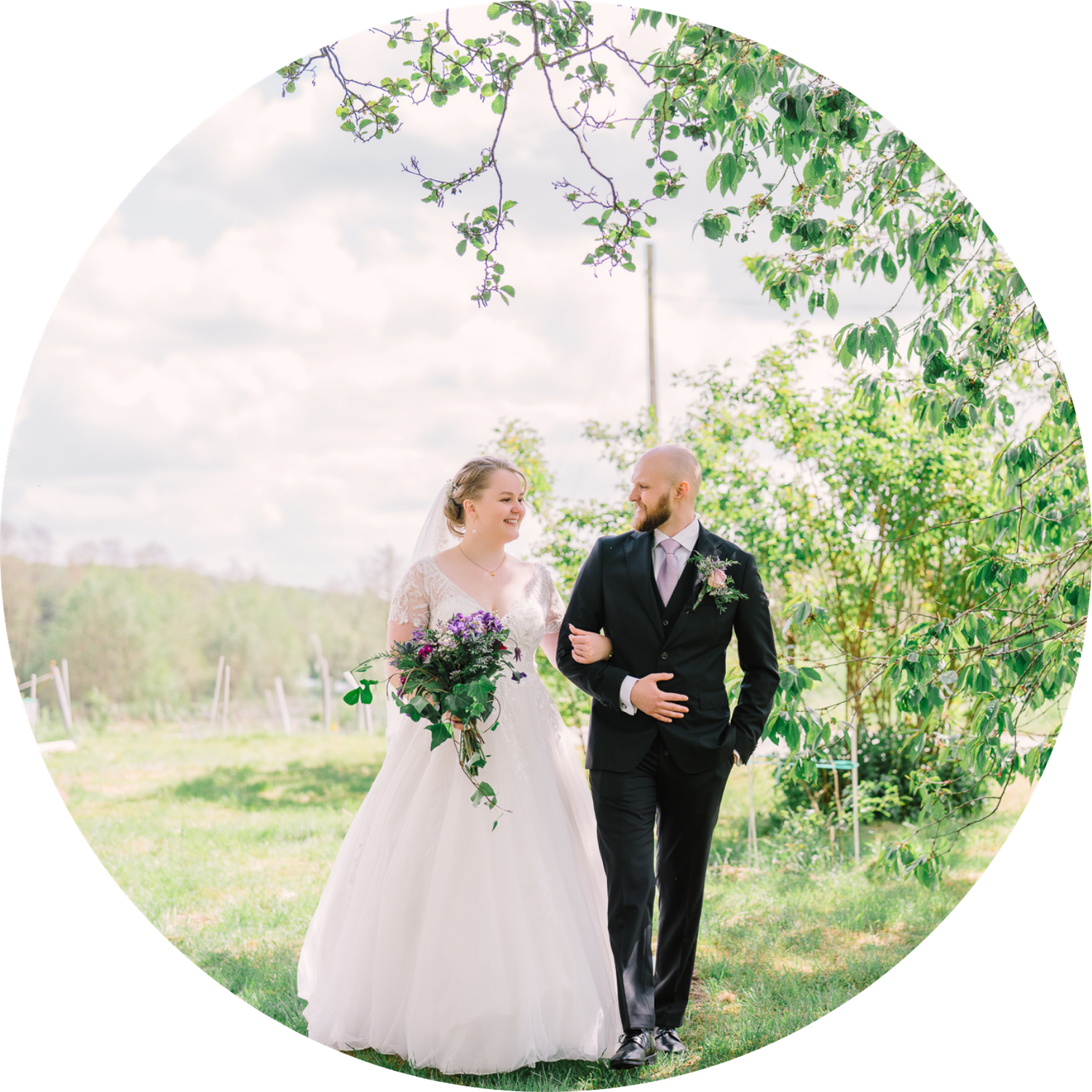 A couple walks arm in arm through a sunlit garden setting on their wedding day in elegant formal attire.