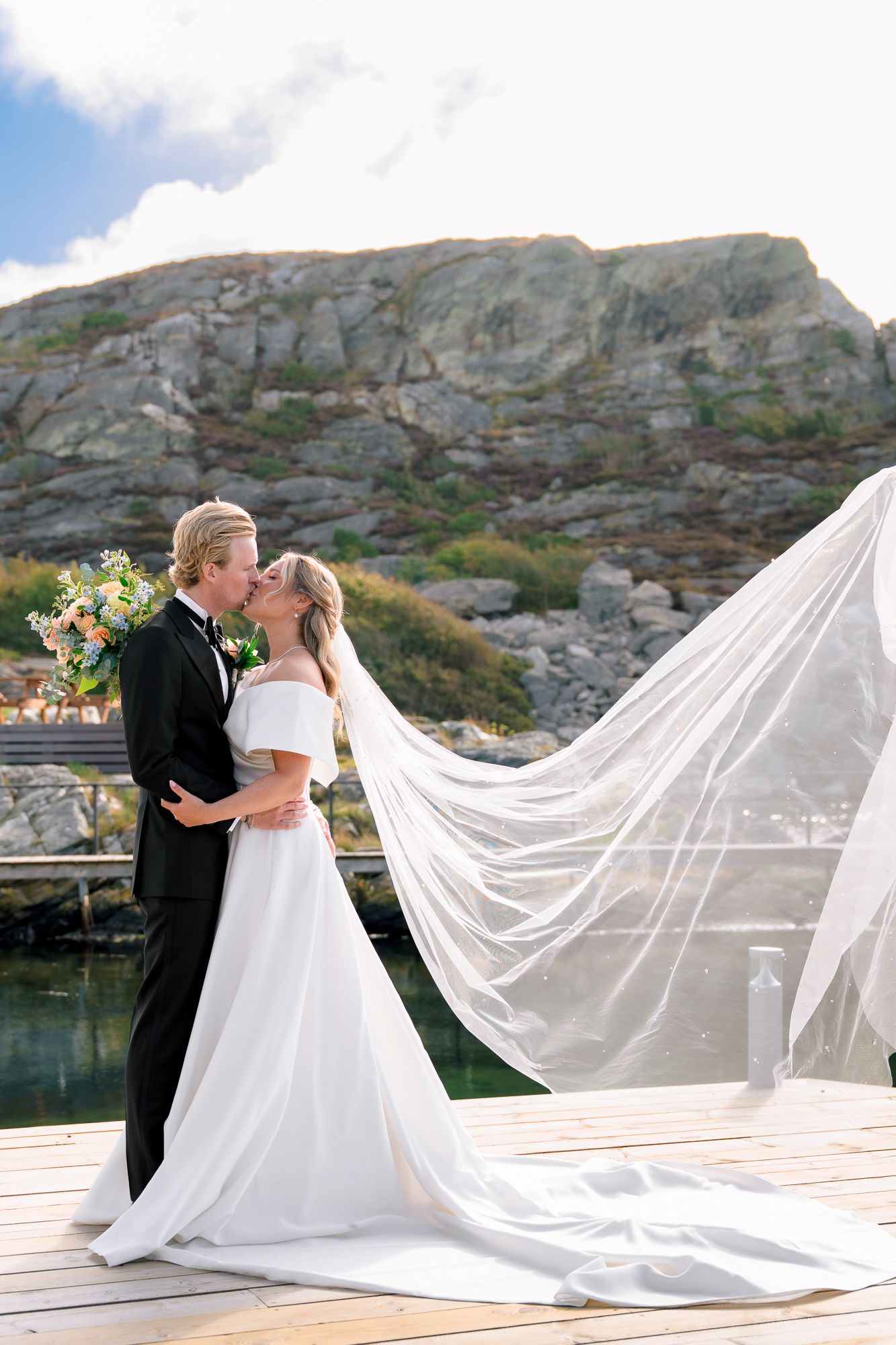 Romantic wedding couple shares a kiss on a dock with flowing white veil and rocky cliffs in the background.