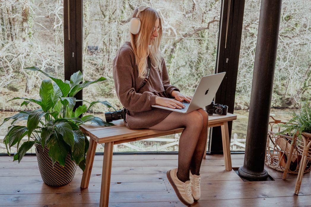 Person working on laptop while sitting on wooden bench with indoor plants nearby.