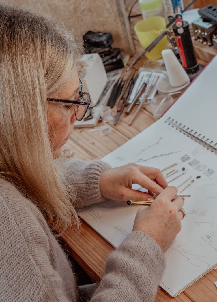 Artist sketching jewellery designs in a notebook at a wooden workspace.
