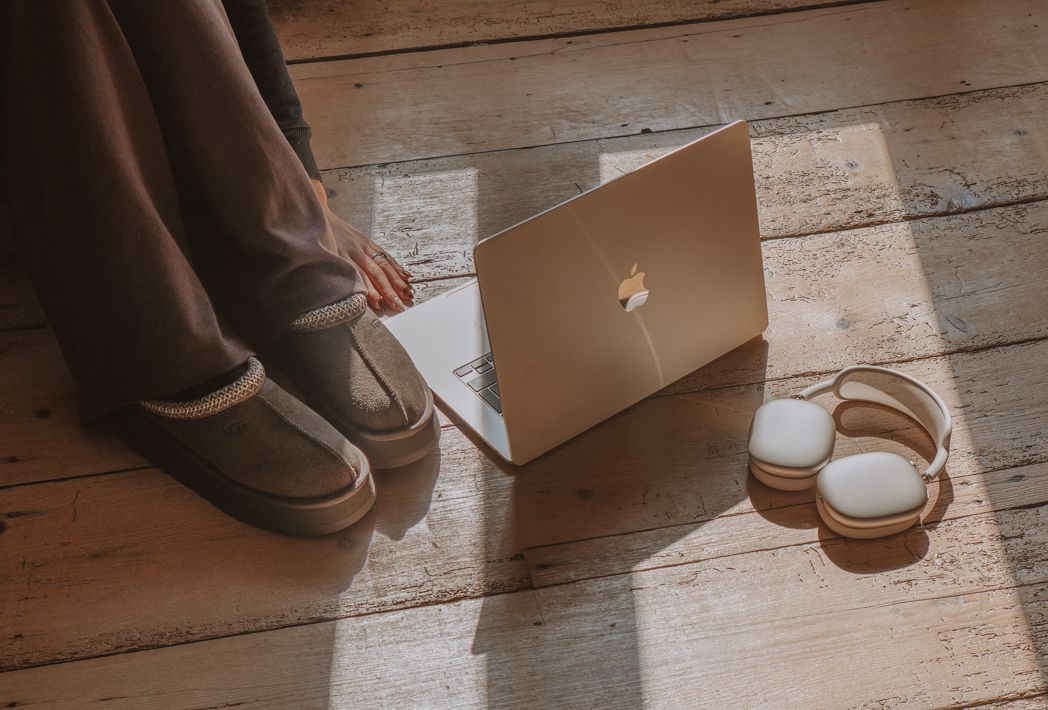 Person wearing brown boots sits on wooden floor with Apple MacBook laptop and headphones in cozy home setting.