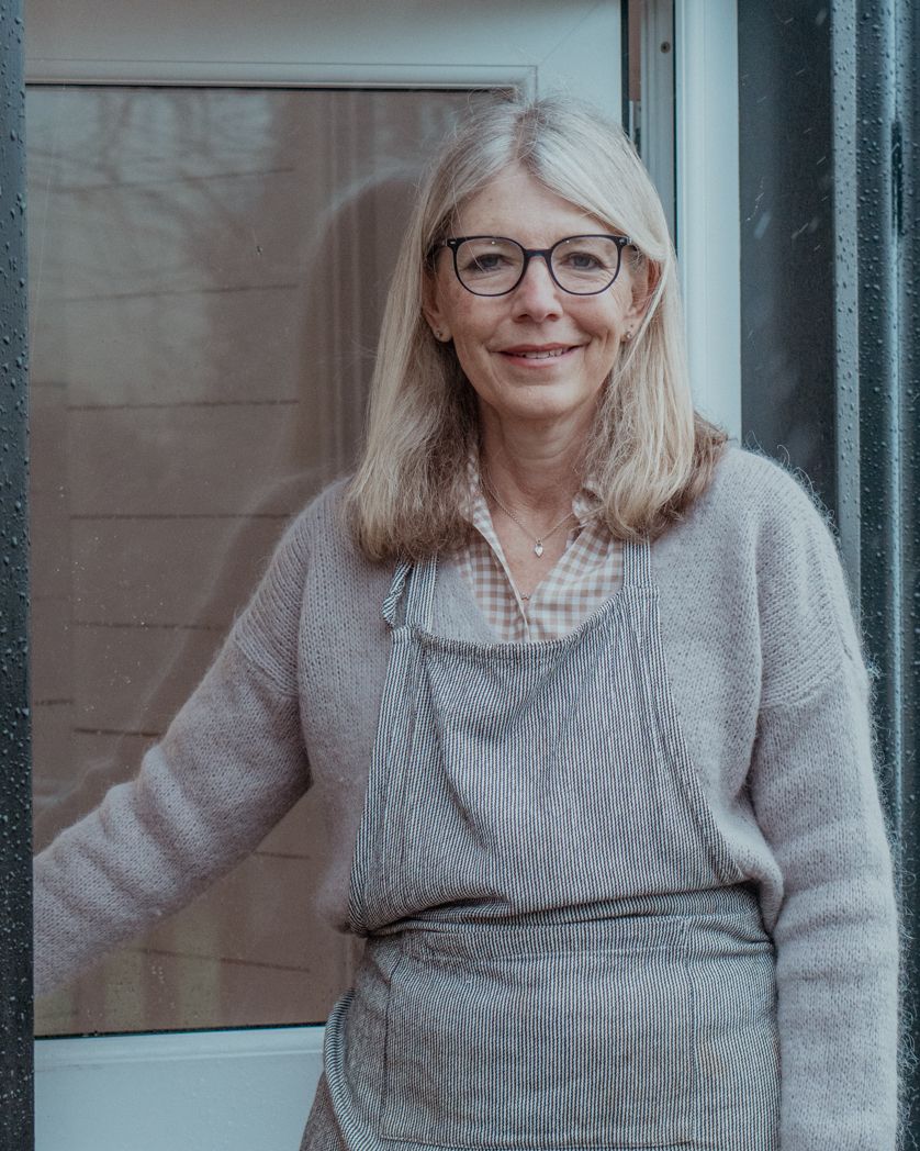 A craftsperson in a gray sweater and apron stands in natural light.