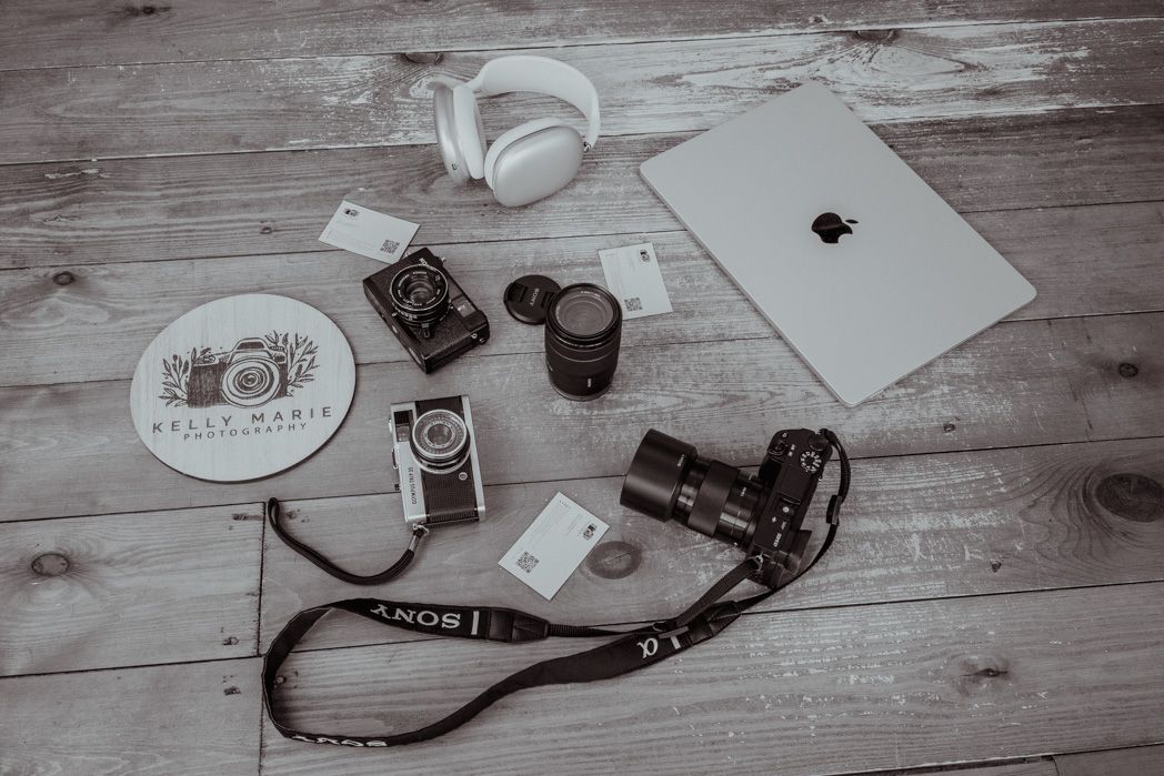 Flatlay of MacBook, camera gear, headphones and coffee on wooden surface.