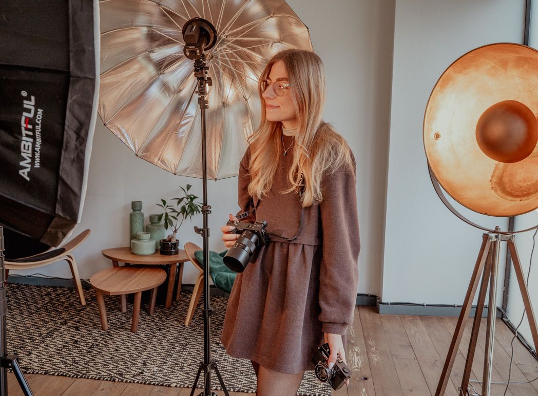 A photographer wearing a brown dress with cameras in a studio setup with professional lighting equipment, reflectors against neutral decor.
