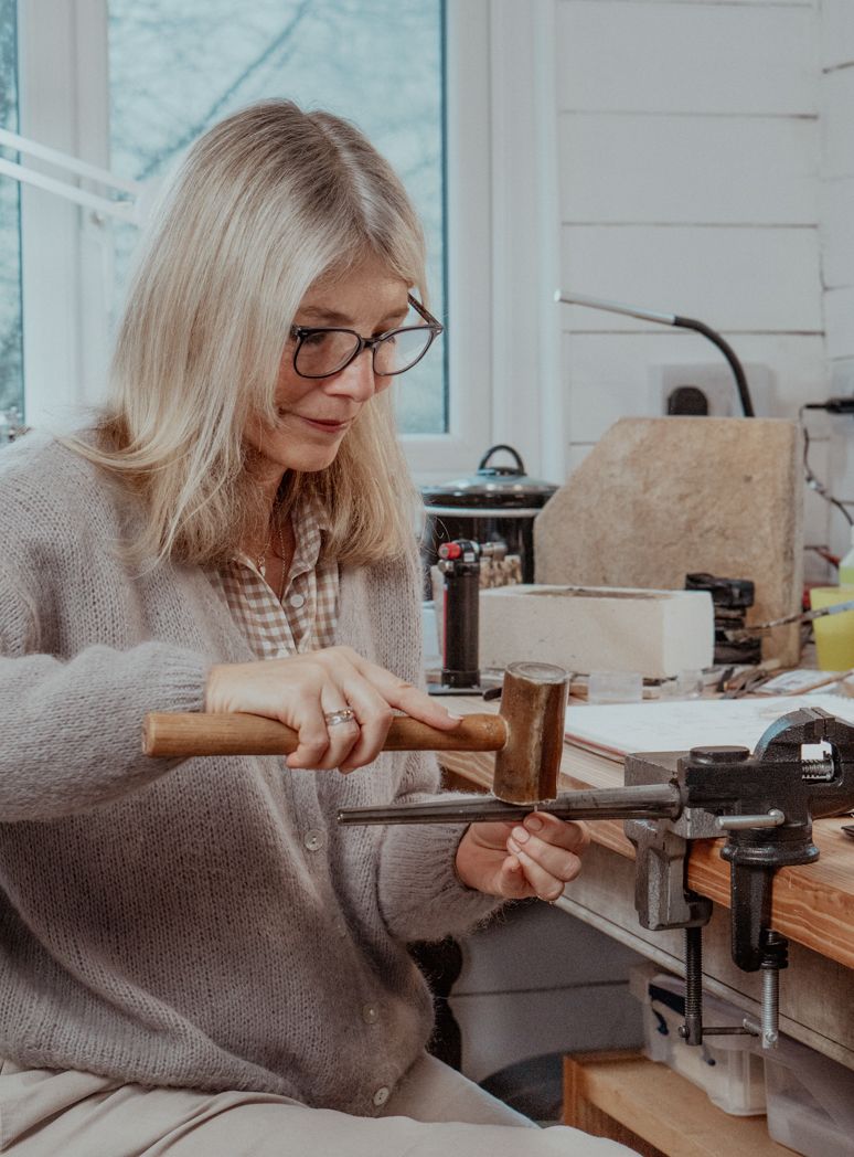 A jewellery uses tools at a workbench while wearing a cosy grey cardigan.