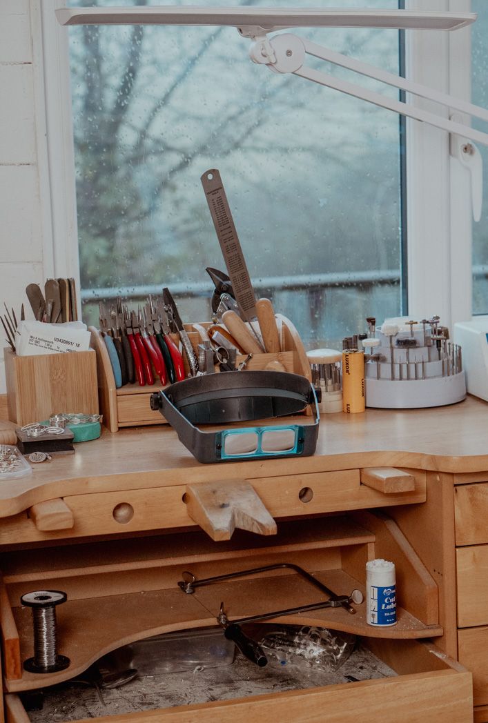 Jewelry making workbench with tools, organizers and magnifying lamp near a window.
