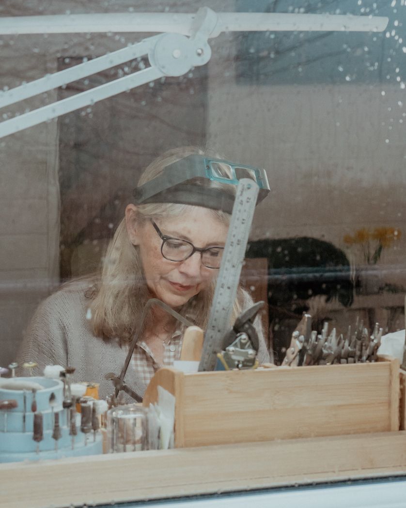 A person working intently at a jewelry or craft workbench with a mounted magnifying lamp and organized tools.