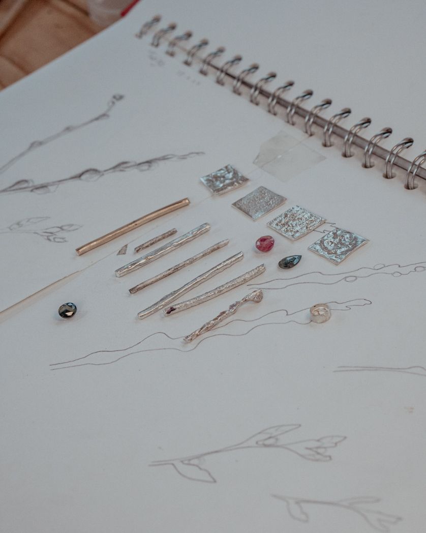 Sketched jewellery designs and tools layout in a spiral notebook on wooden surface.