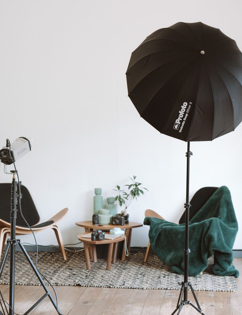 Photography studio setup with black umbrella light, tripod, and emerald green armchair against white wall.