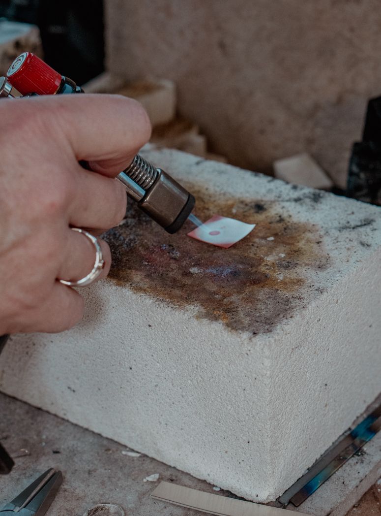 Hand using a metalworking tool on a white surface in a jewellerystudio.