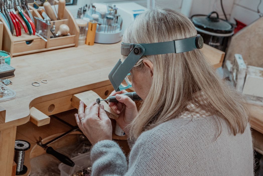 A jewelry maker wearing a magnifying headpiece works on detailed metalwork at a wooden crafting bench.