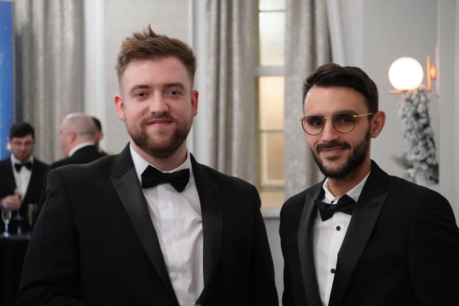 Two men in black tuxedos and bow ties stand together at a formal event with curtains and sconces in background.