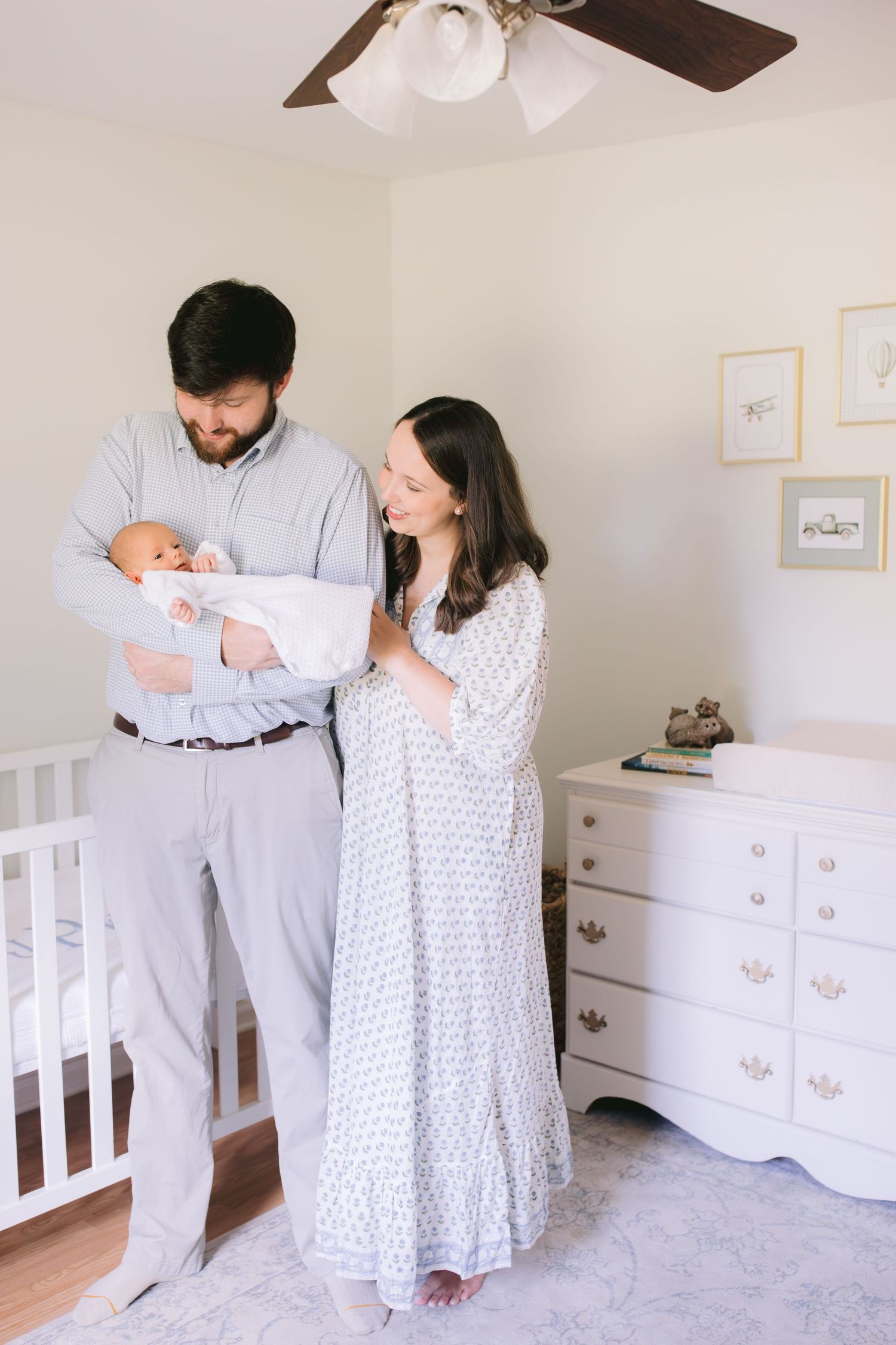 Parents in gray and white outfits share tender moments with their newborn baby in a serene nursery setting.