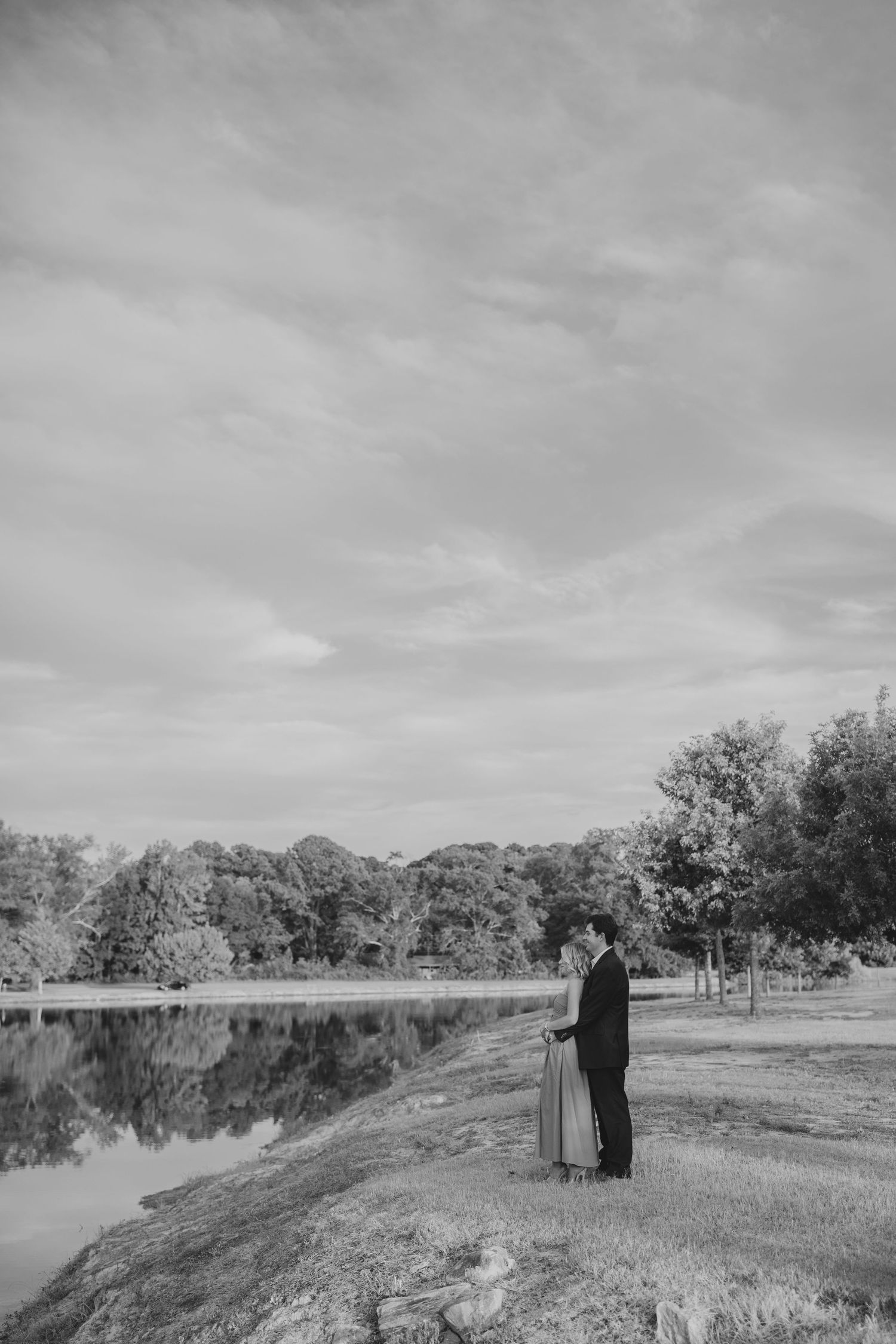 Black and white photograph of a couple standing together in a serene natural landscape beside a tranquil lake.