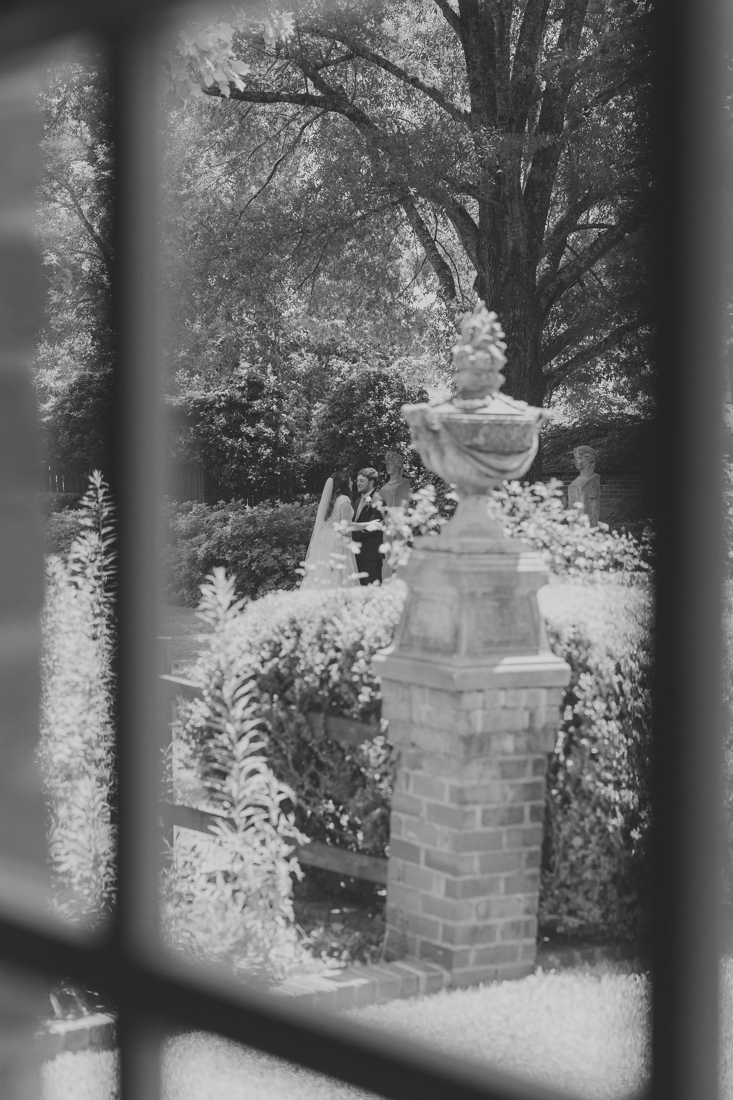 Black and white view through window bars of brick pillars and garden foliage.