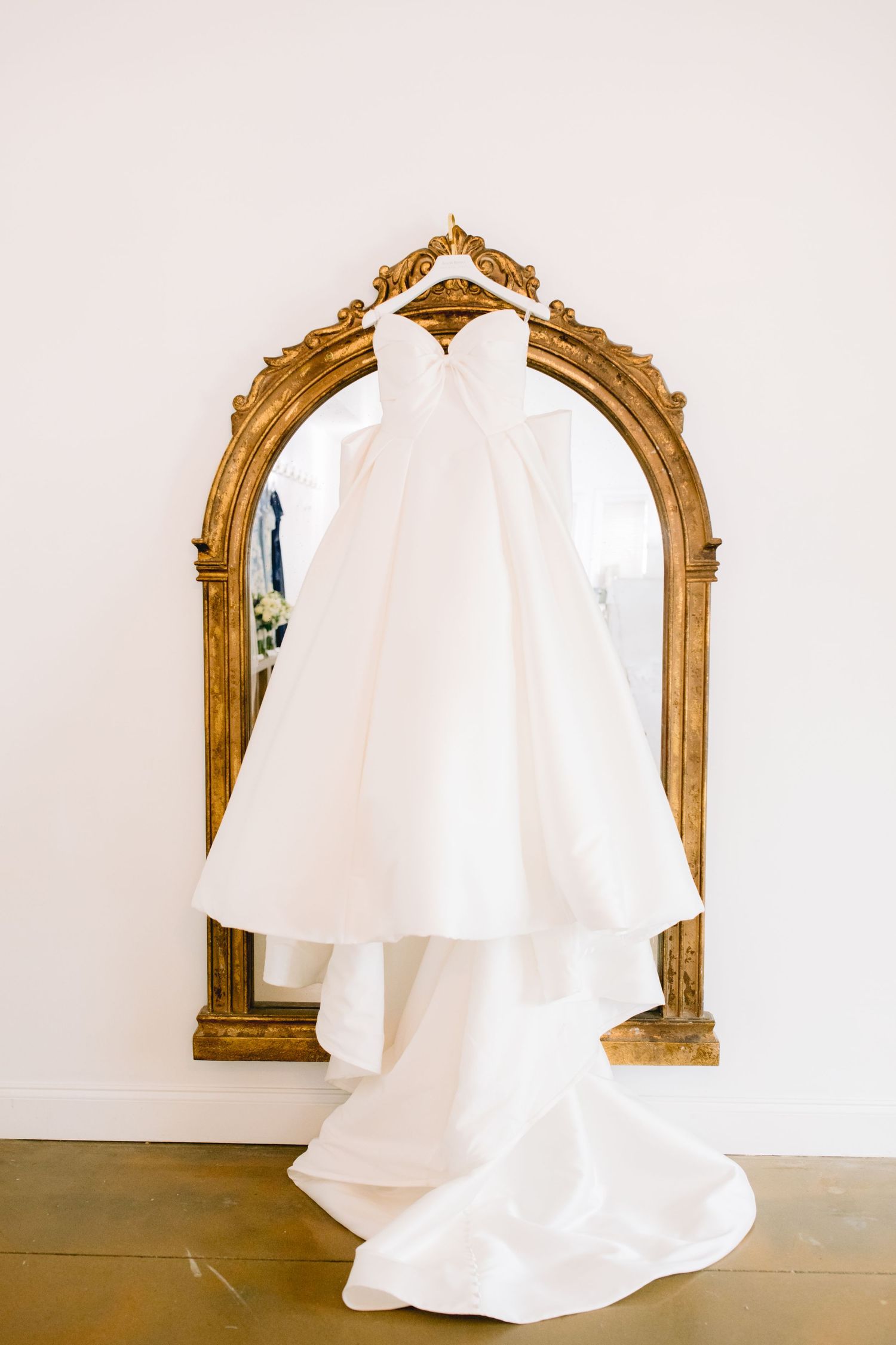 White wedding dress displayed on an ornate gold-framed mirror against a neutral wall.