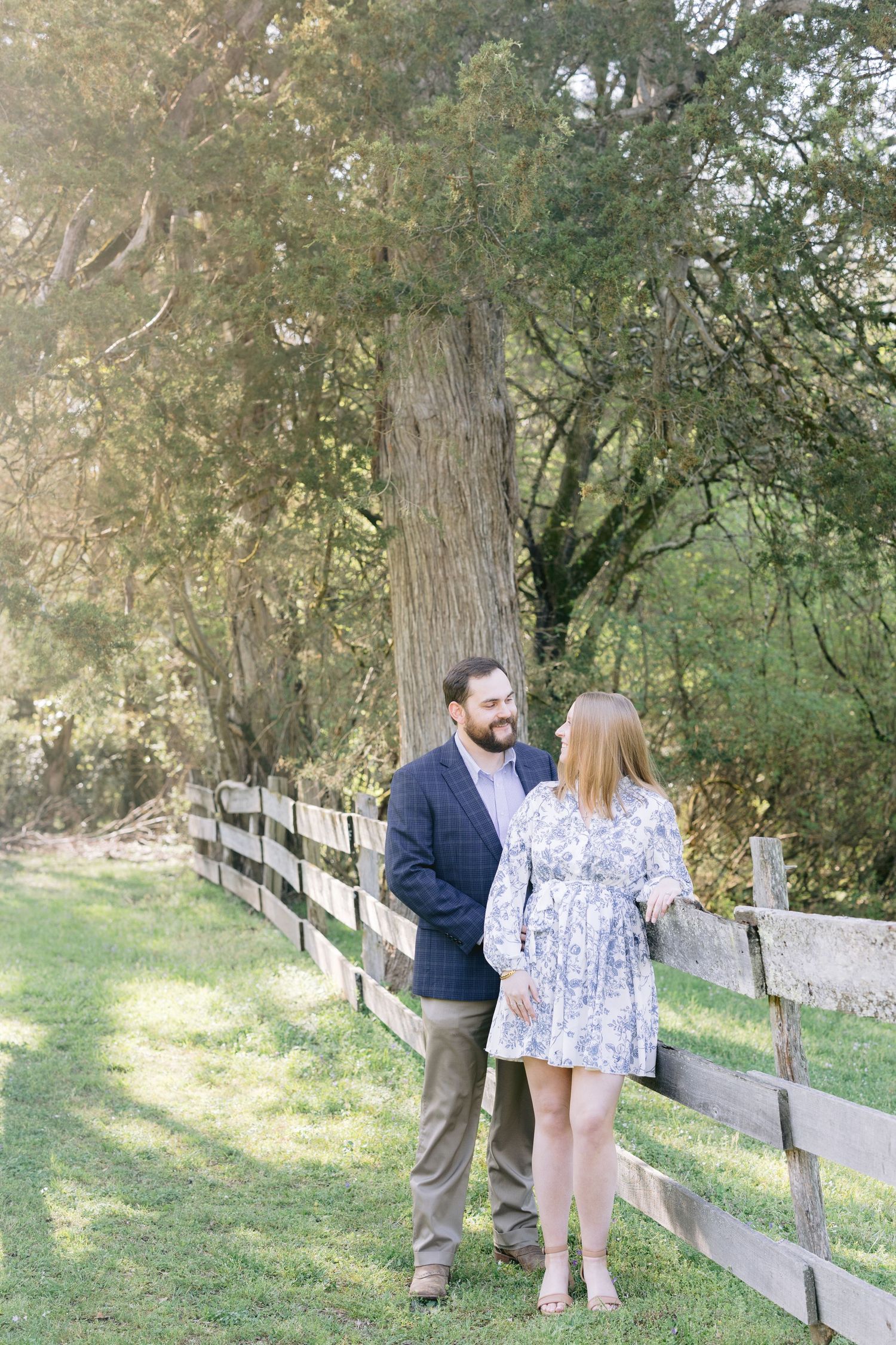 A couple in coordinated outfits sharing romantic moments along a wooden fence path surrounded by trees.