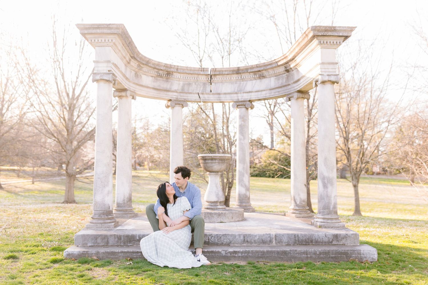 A couple embraces during their wedding photo session in a white stone gazebo surrounded by lush green grass.