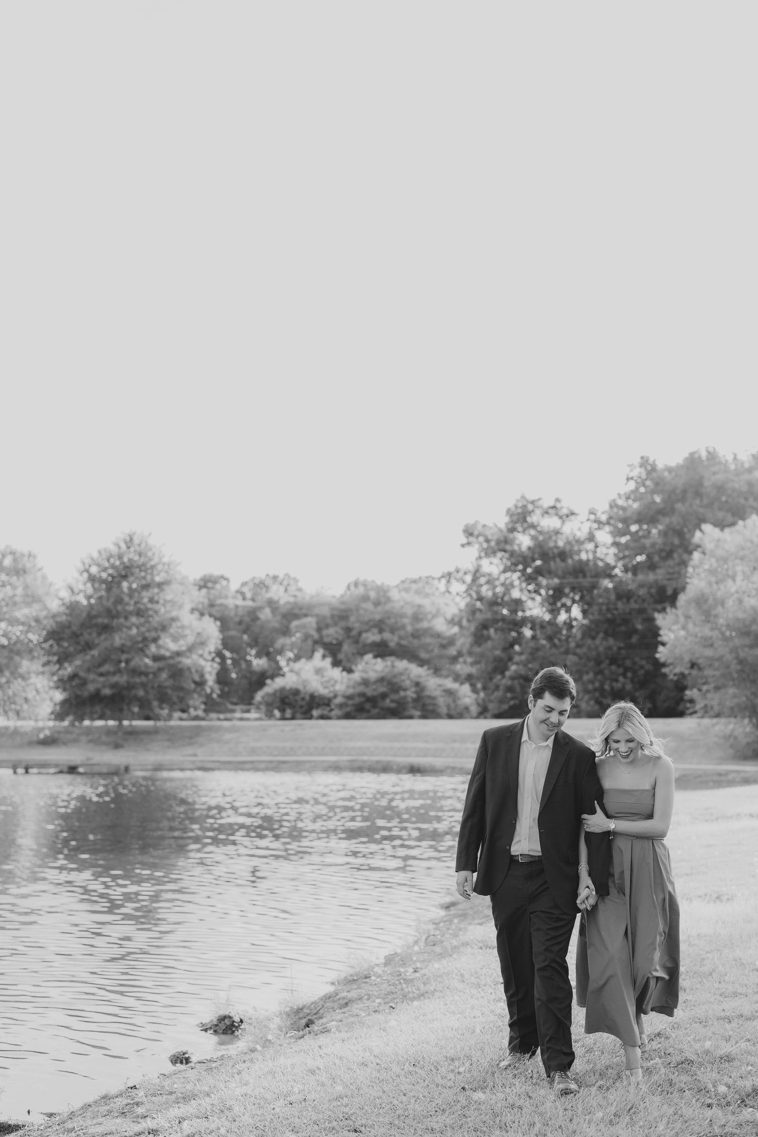 A couple walks hand in hand beside a serene lake on an overcast day.