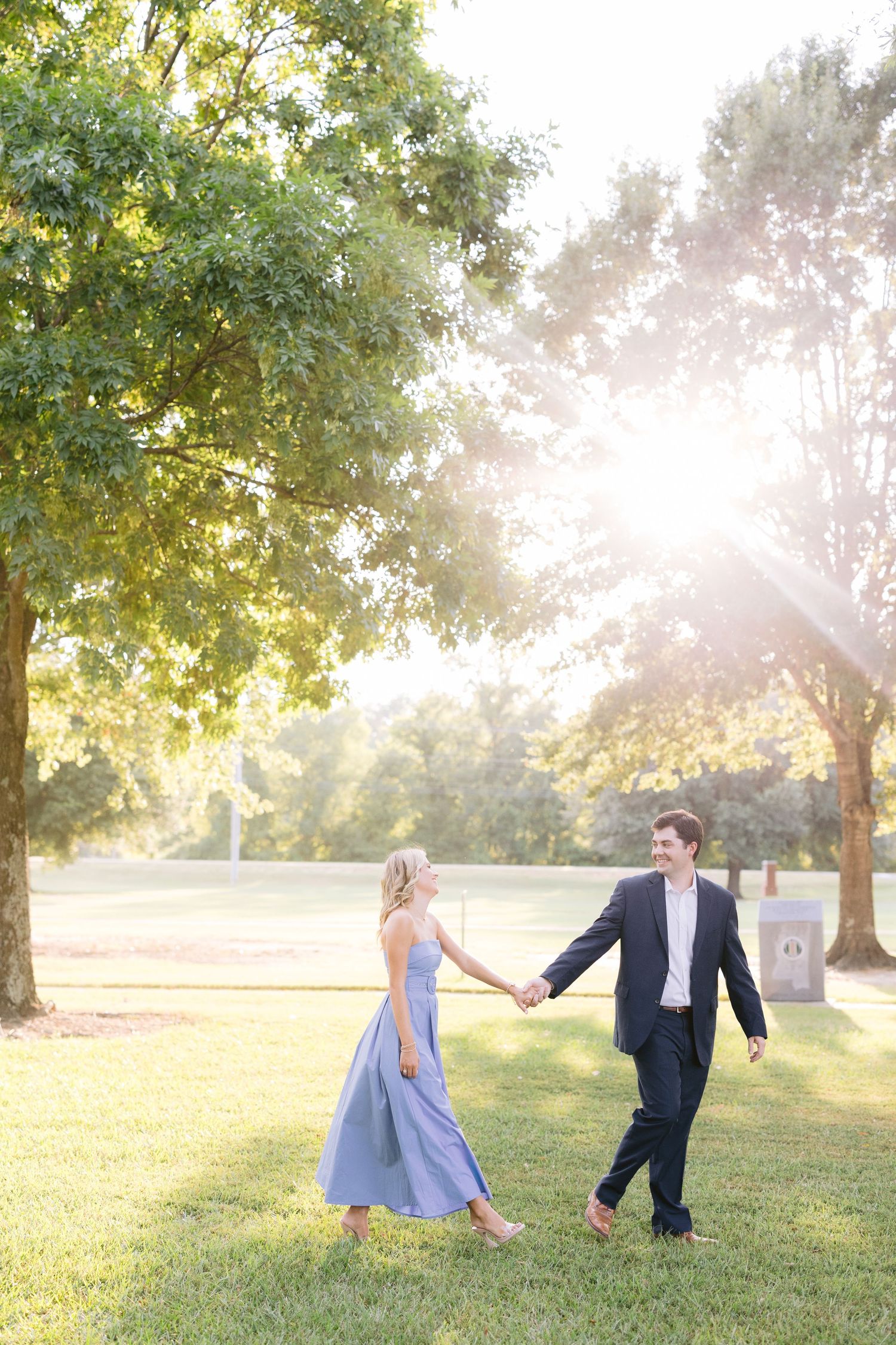 A couple walks hand in hand through a sun-drenched park with golden light filtering through trees in their formal attire.