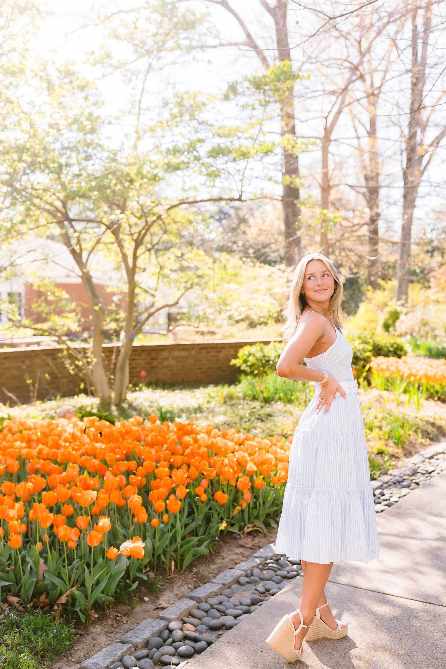 Young woman in white sundress poses gracefully among rows of vibrant orange tulips on a garden path.