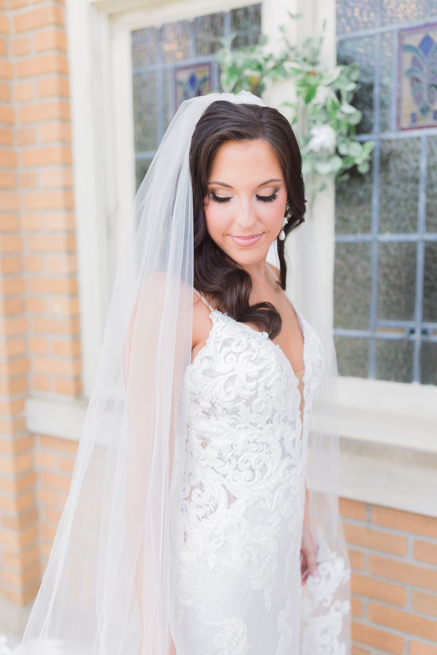 Bride in lace wedding gown with veil poses by brick window in natural light.