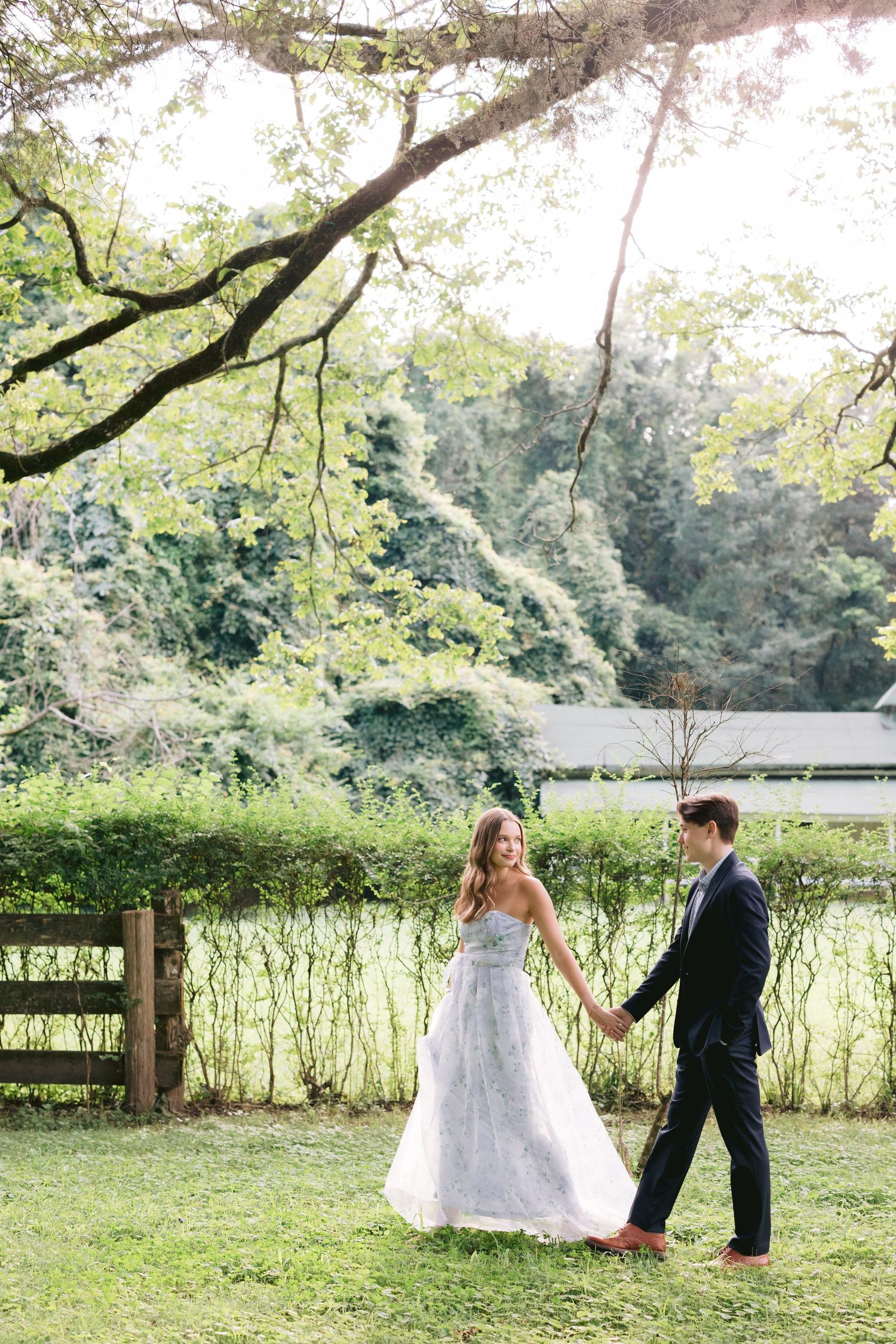 Bride and groom walking together through a lush garden setting with green foliage and overhanging trees in sunlight.