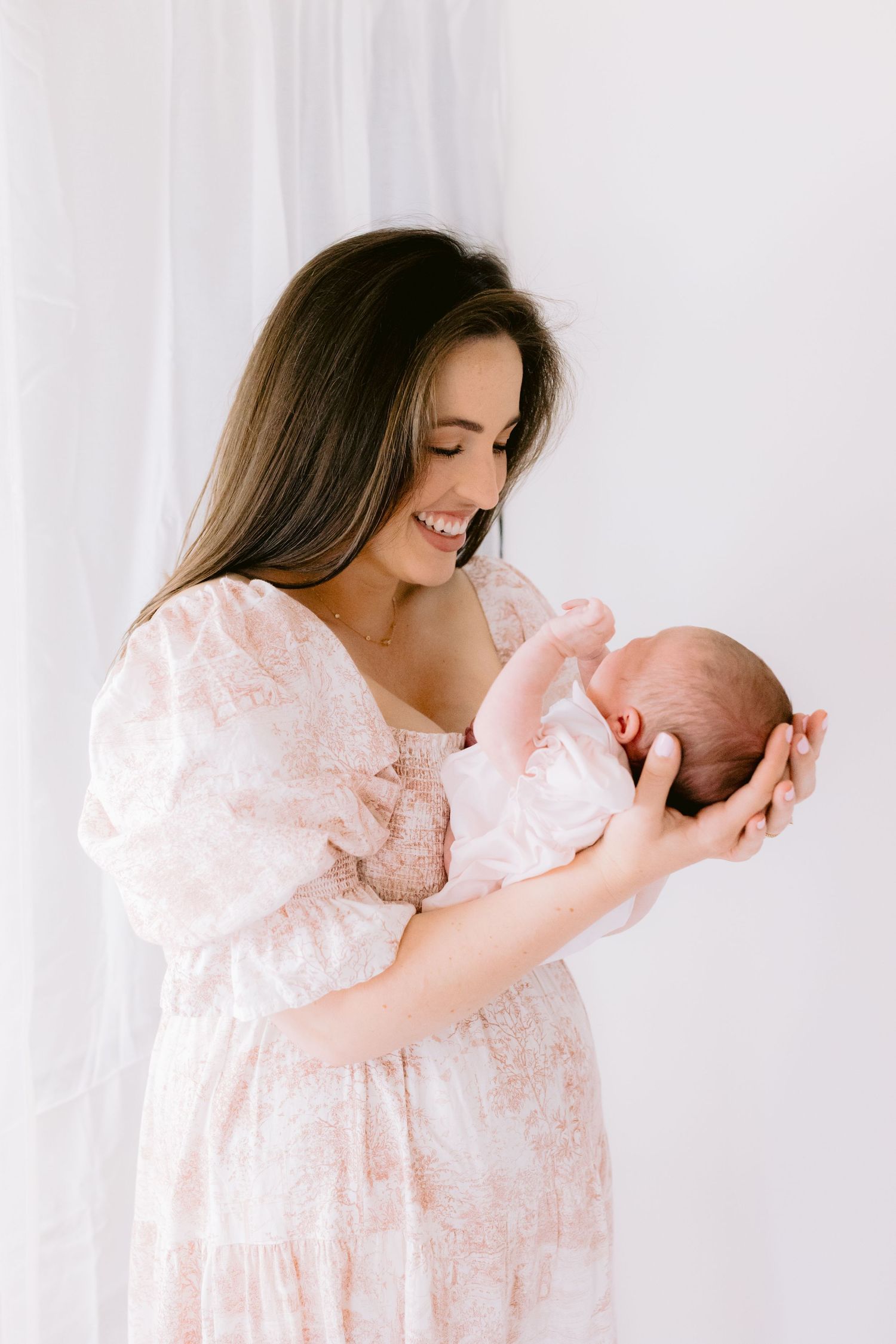 A parent in pink floral dress holds and snuggles with a newborn baby in a bright white studio setting.