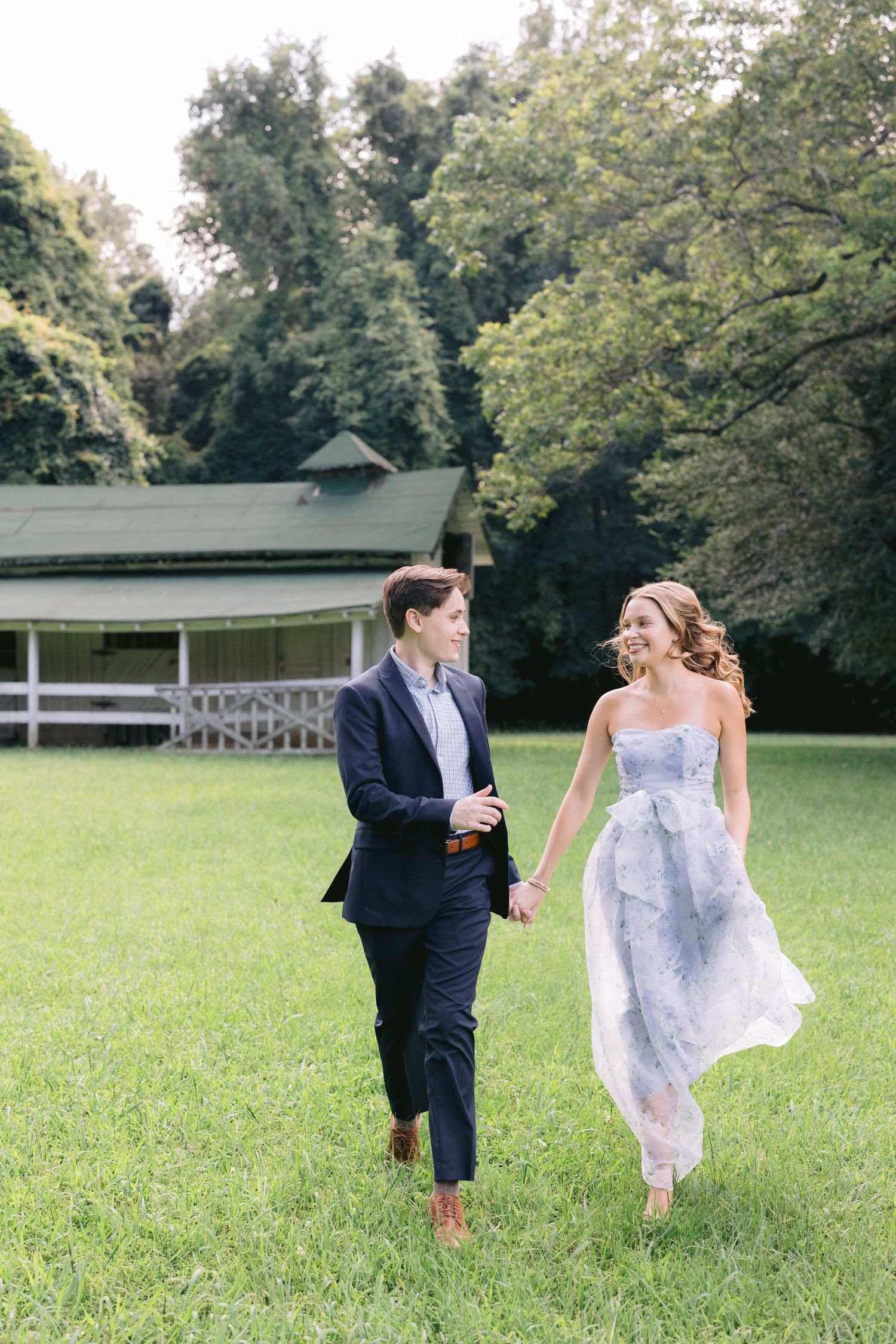 A couple playfully runs and chases each other across a lush green field during an outdoor photoshoot.