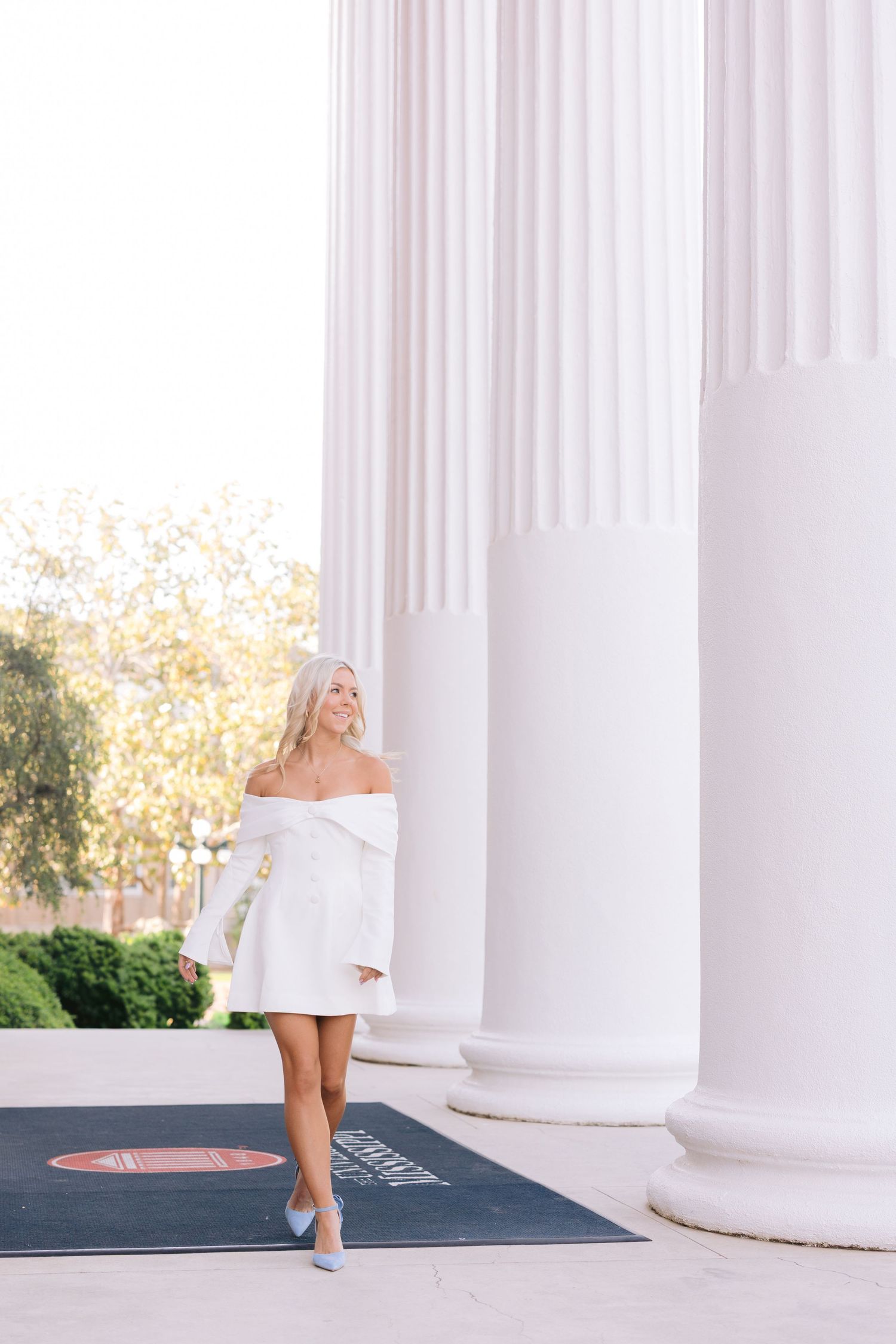 Woman in white off-shoulder dress poses elegantly between classic white columns in an outdoor setting.