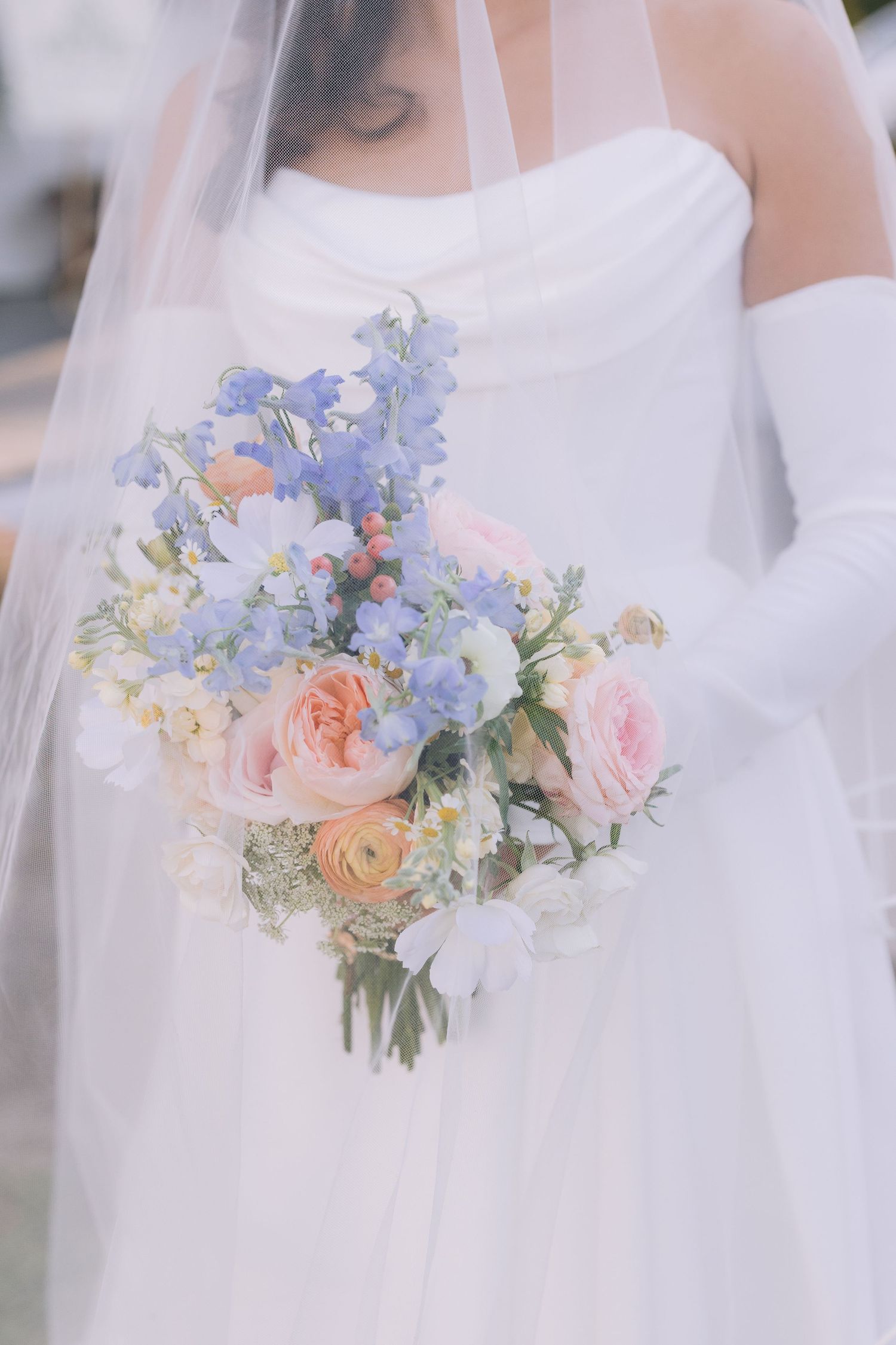 Delicate pastel wedding bouquet featuring blue and peach flowers against white dress.