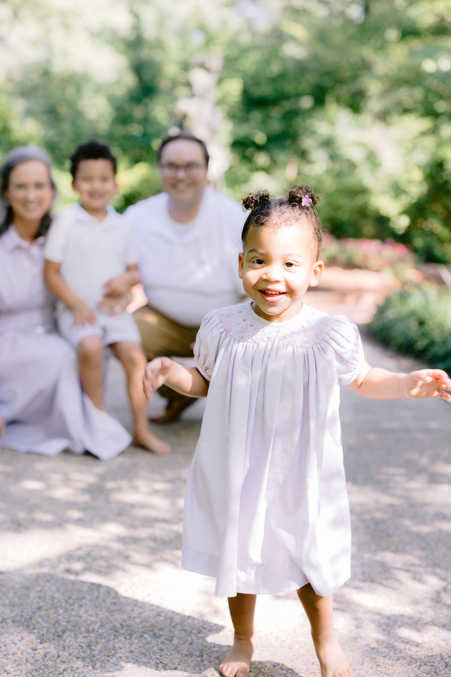A toddler in a white dress stands and smiles while playing outside on a gravel path on a sunny day.