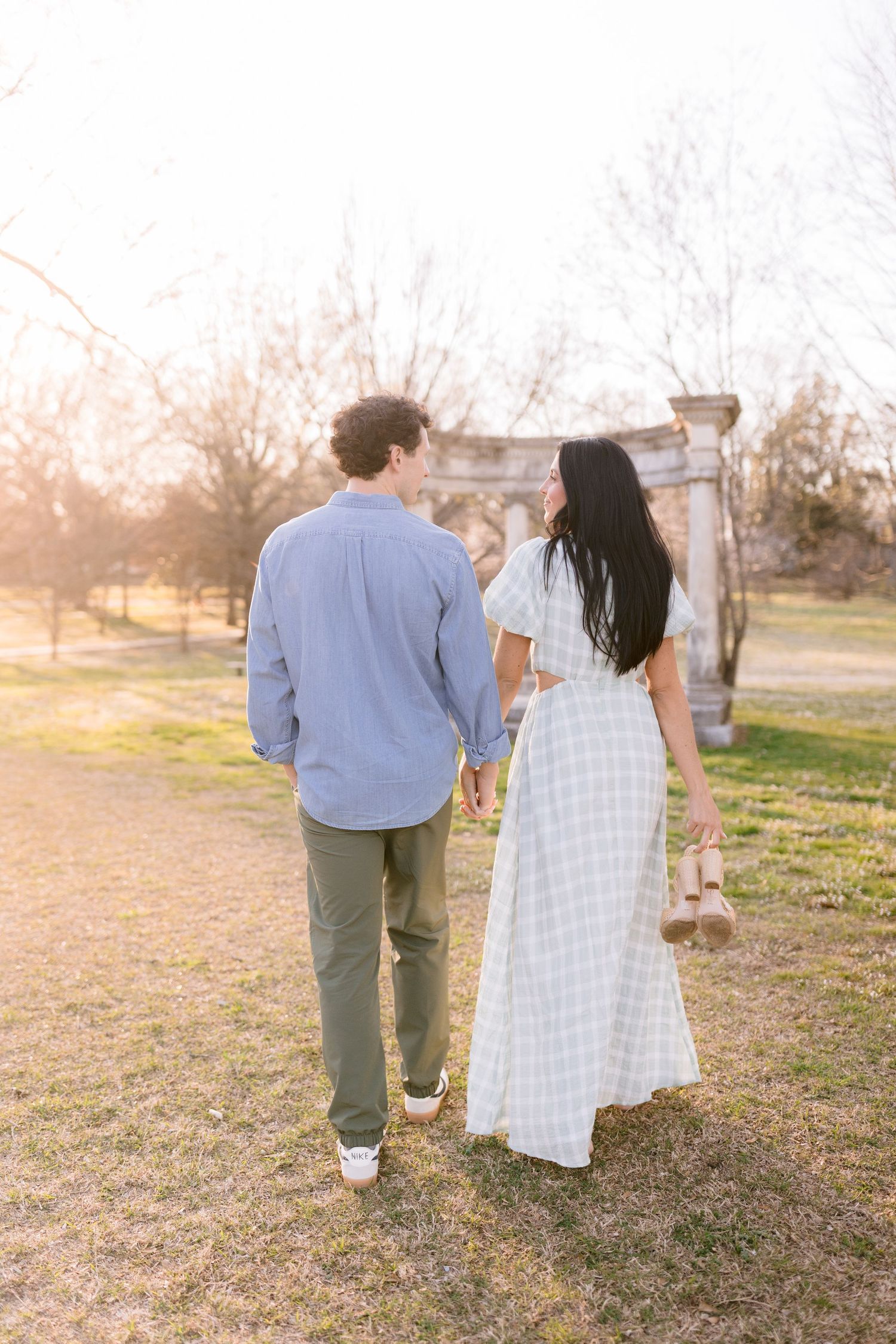 A couple walking hand in hand through a sunlit field at sunset, photographed from behind in romantic poses.