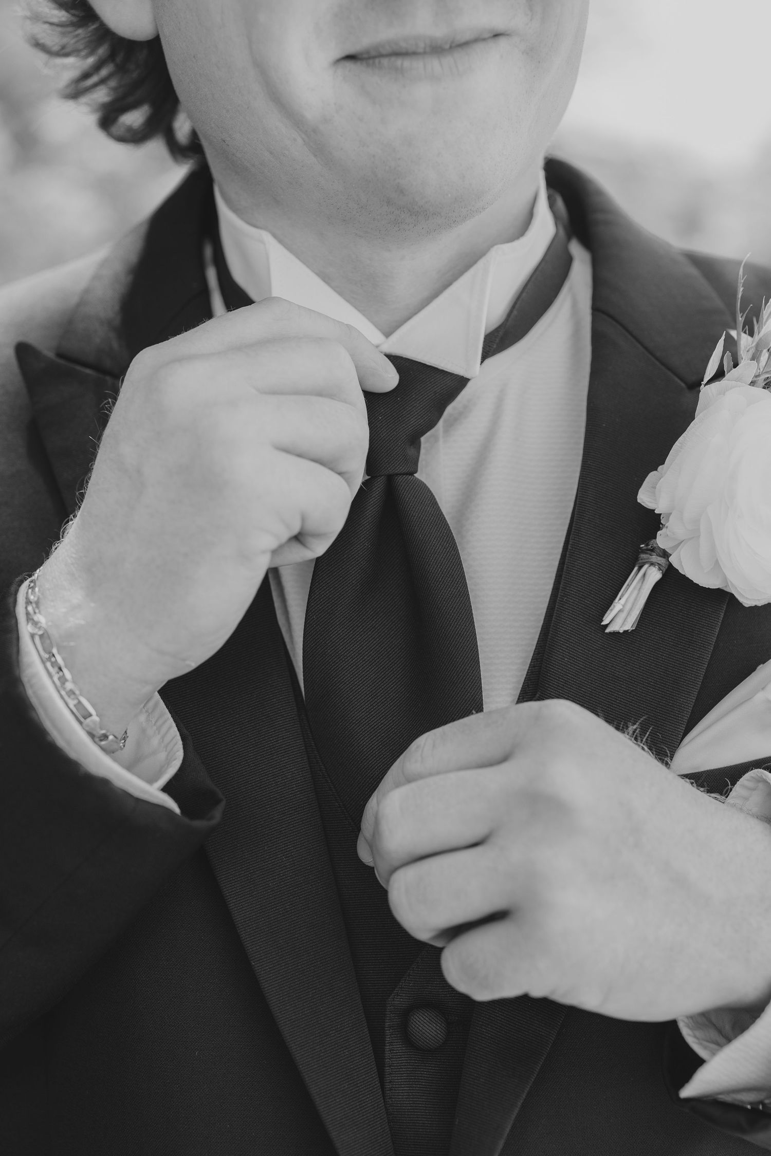 Close-up black and white portrait details of groom adjusting tie with boutonniere.